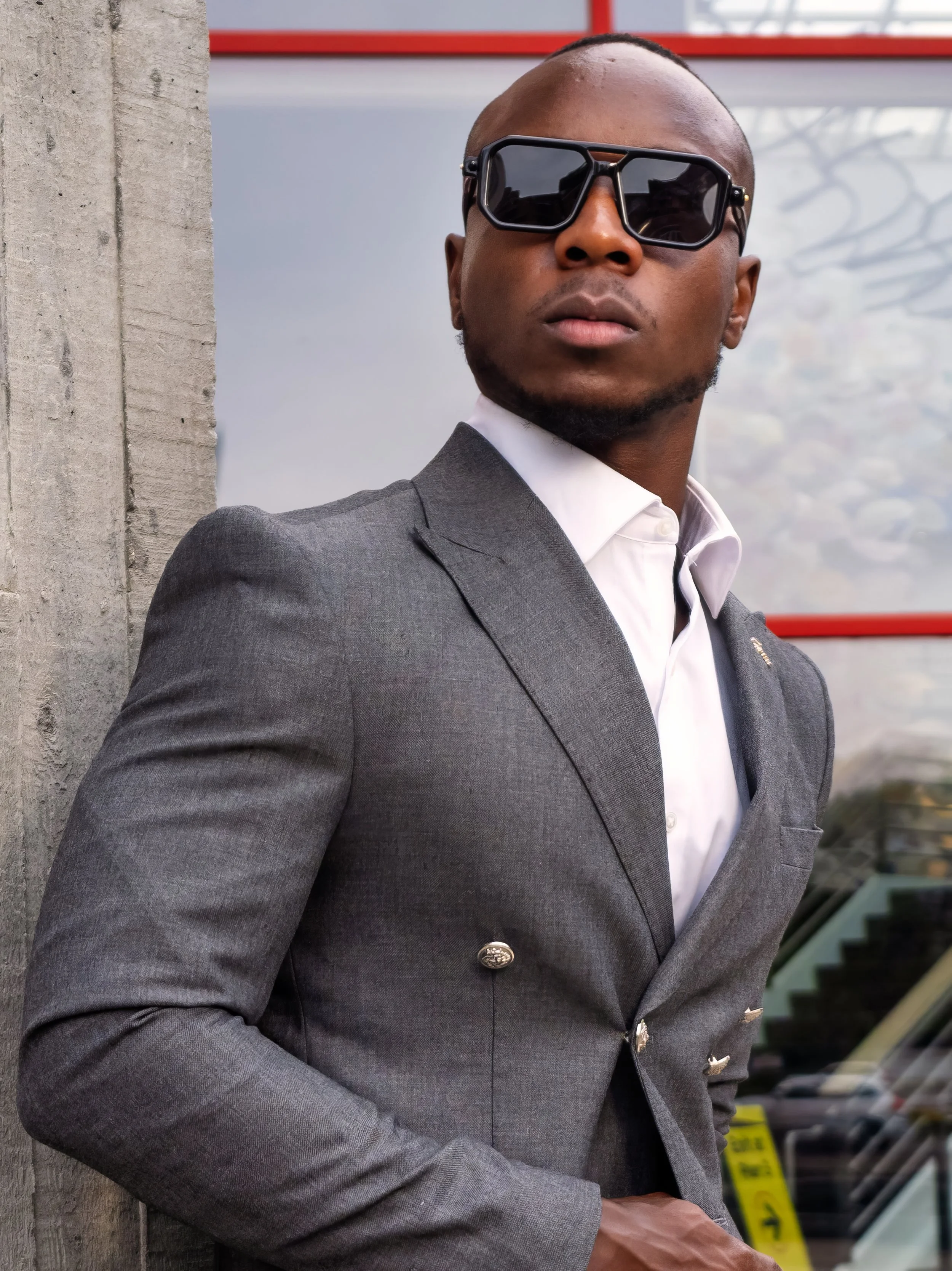 A man wearing a gray suit, white shirt, and large black sunglasses standing outdoors beside a concrete wall with a cityscape reflected in the background window.