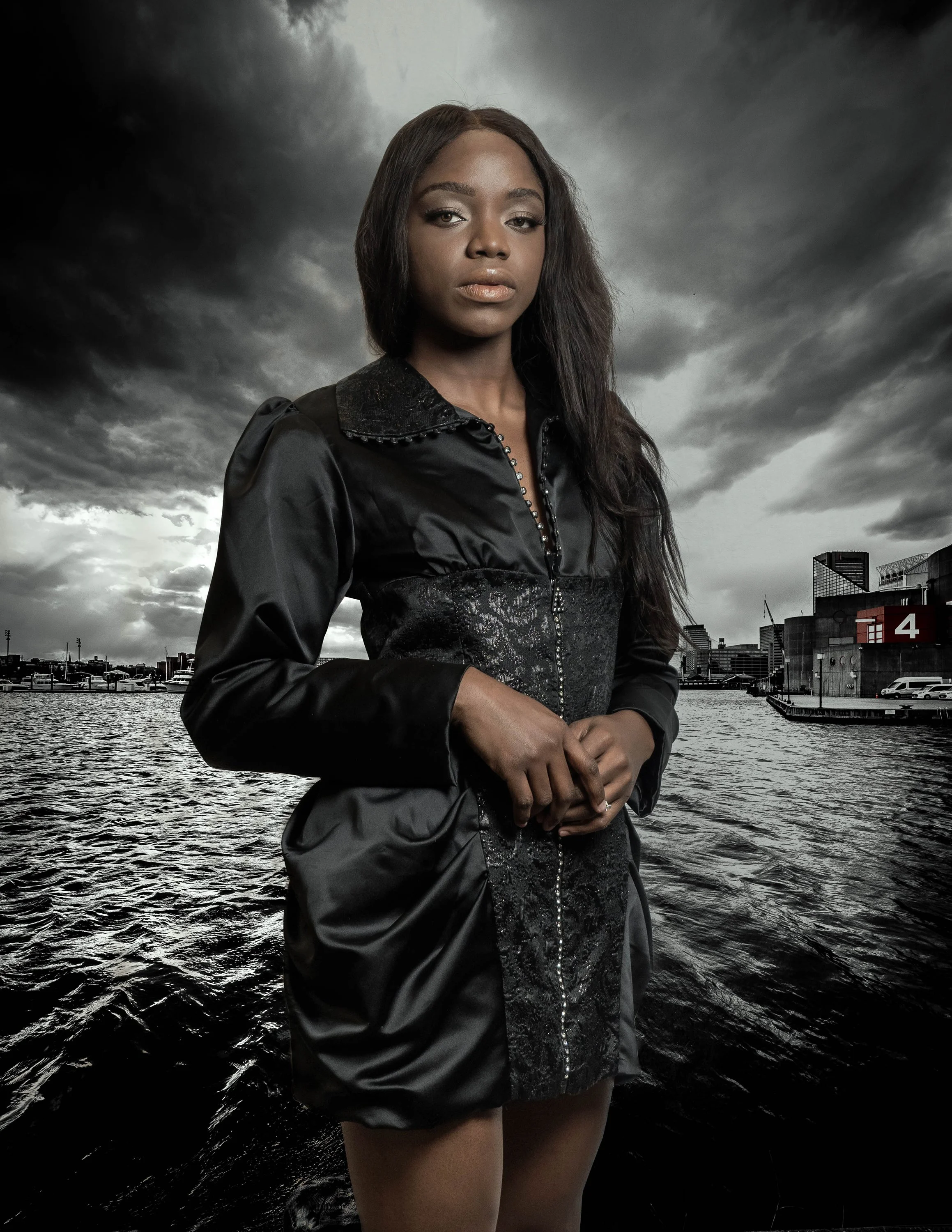 A woman with dark skin and long dark hair standing near a body of water under dark stormy clouds, wearing a black shiny dress with a zipper and lace details.