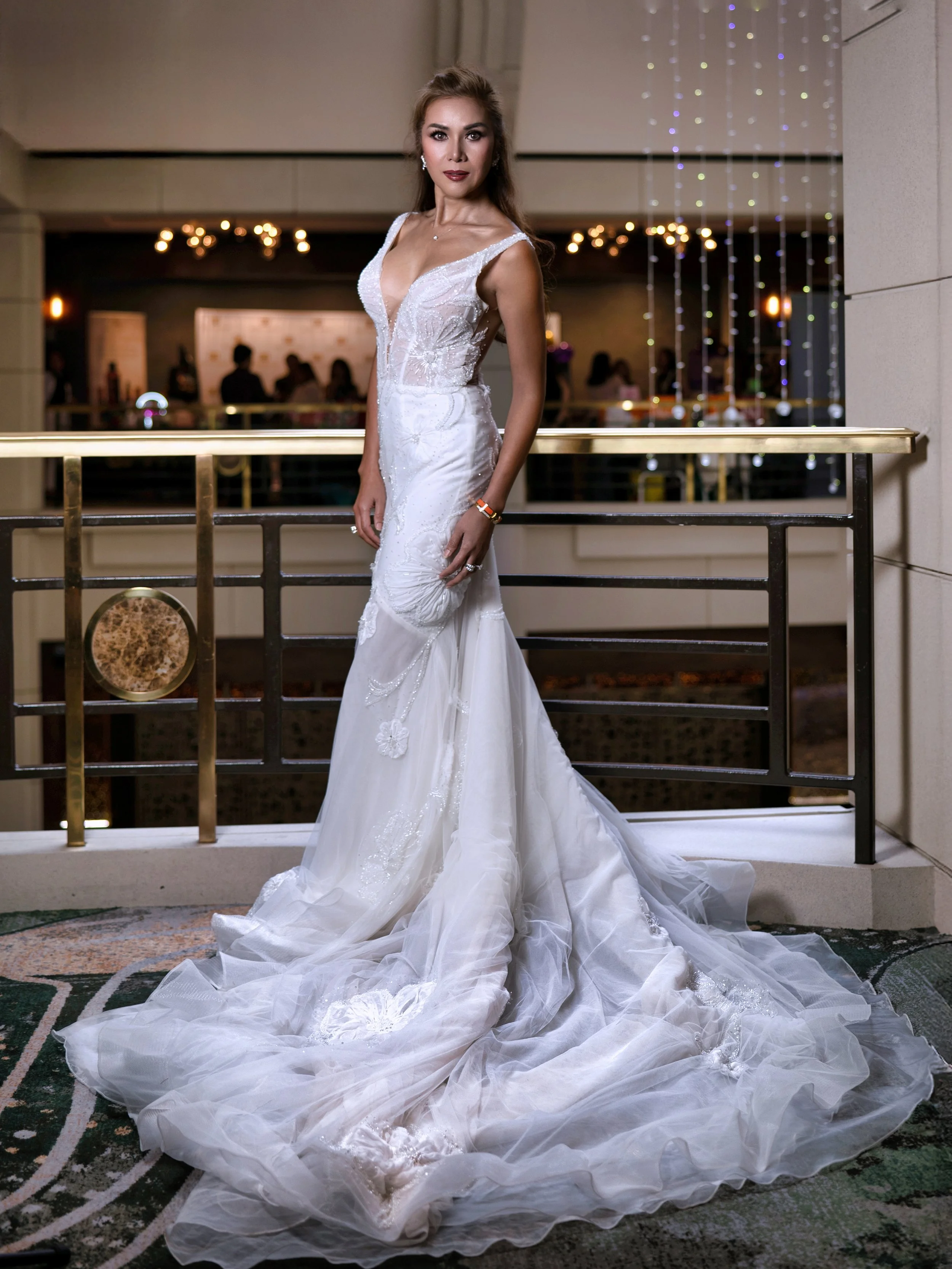 A woman in a white wedding dress standing on a balcony with a decorative railing, inside a venue with warm lighting and people in the background.
