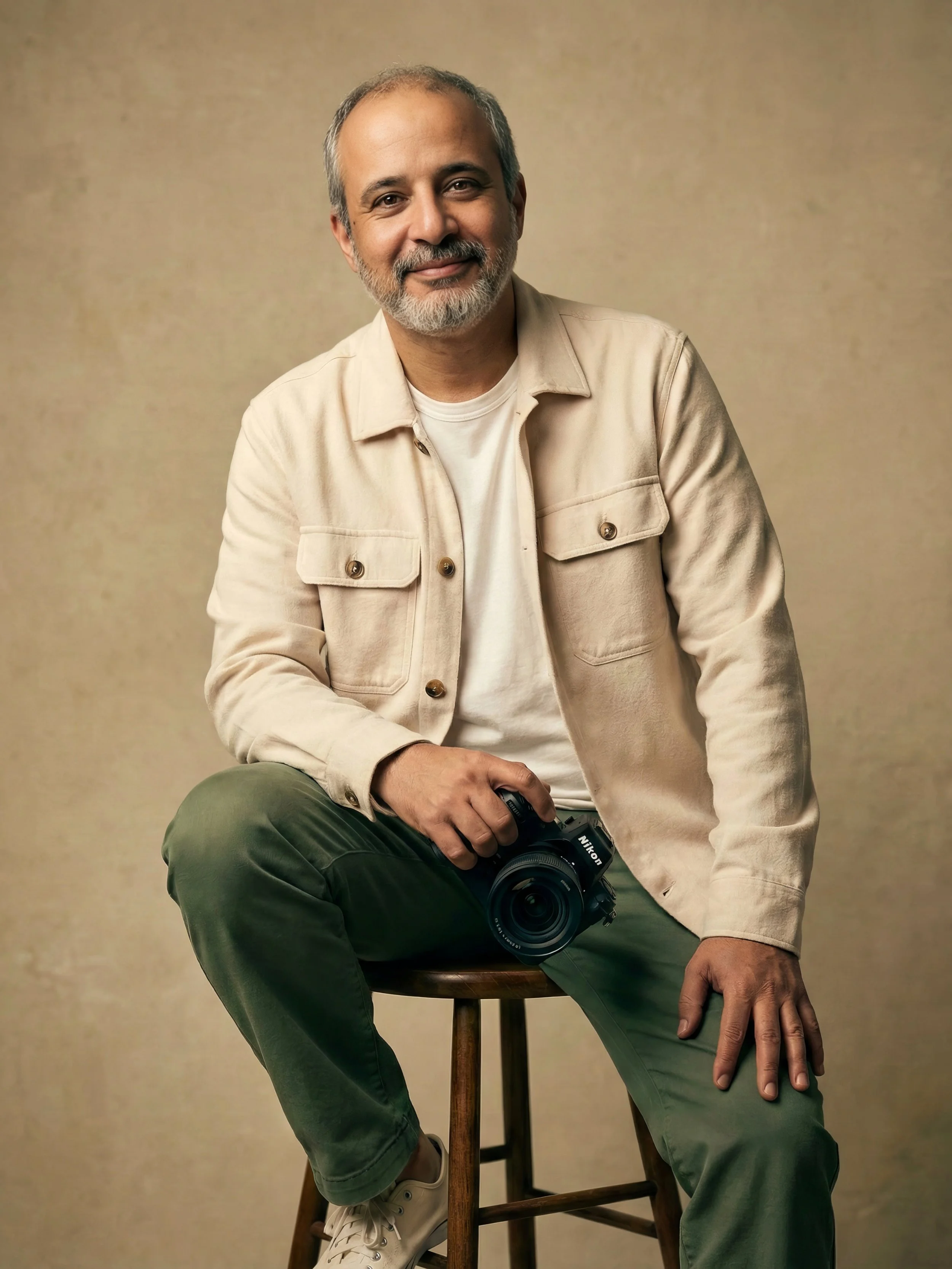 A middle-aged man with gray hair and a beard, wearing a beige jacket, white shirt, and green pants, sitting on a wooden stool holding a Nikon camera, smiling at the camera.