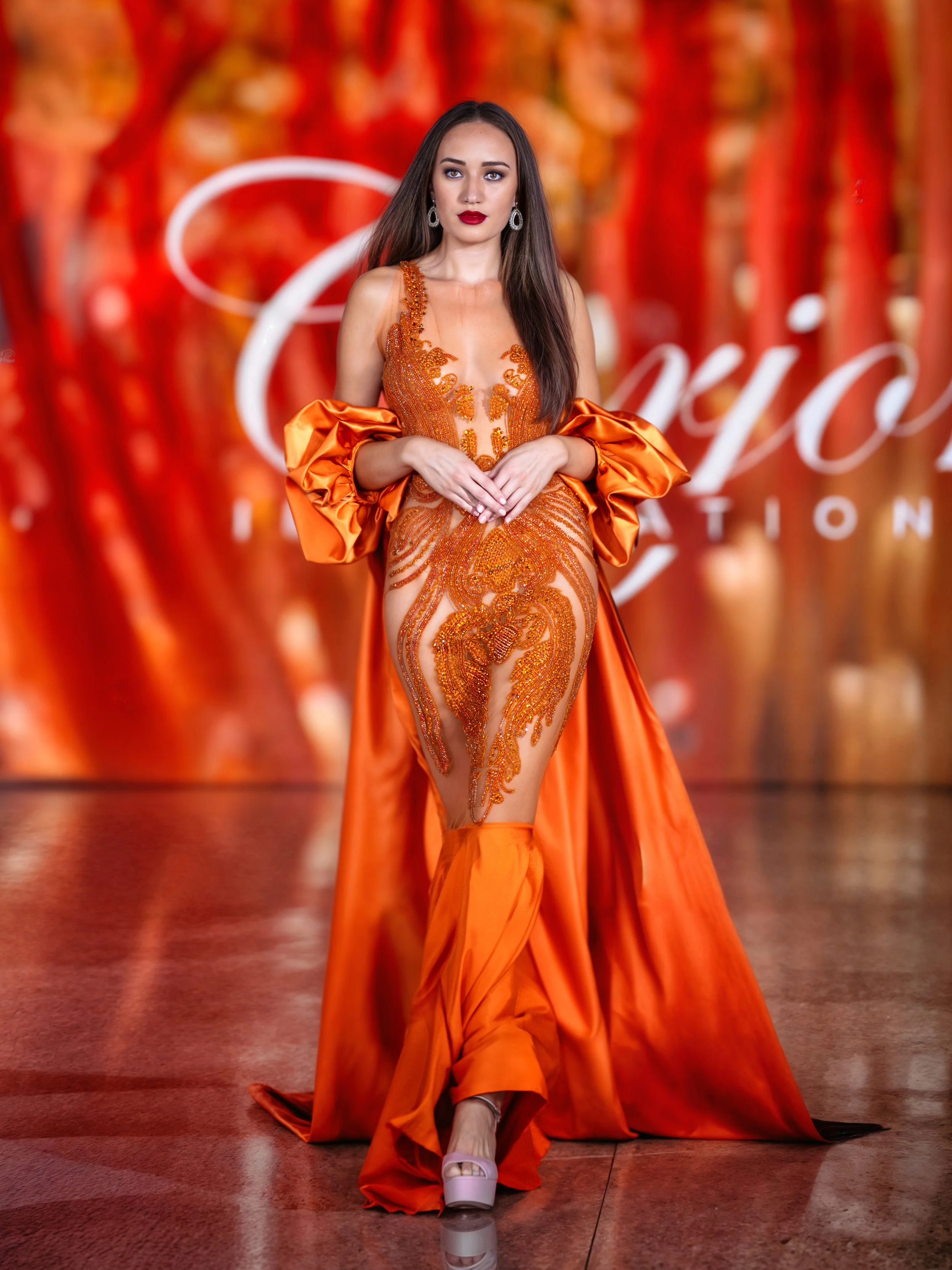A female model walking on a runway in an elaborate orange gown with intricate embroidery and sheer panels. The gown has a long train, puffy sleeves, and a dramatic skirt with high slit, paired with high heels. The background features a blurred red ba