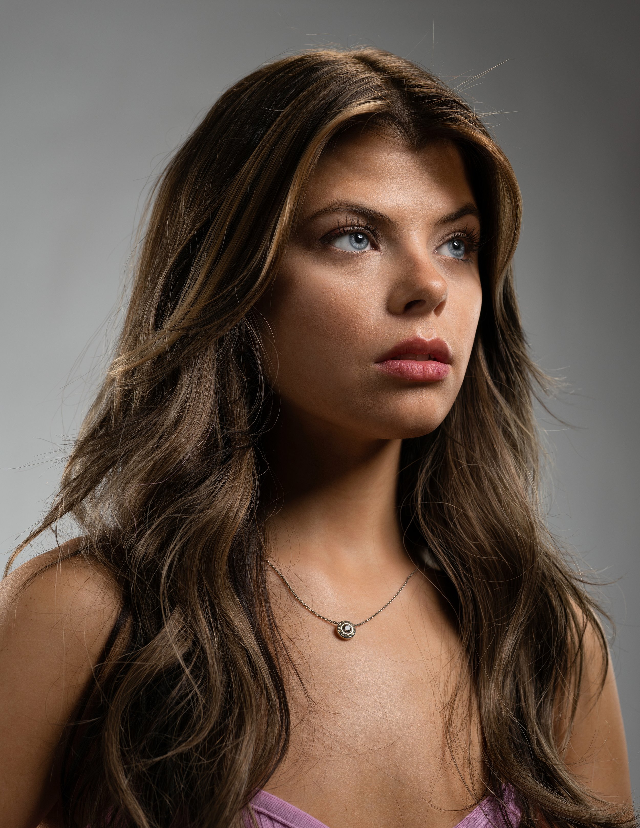 Portrait of a young woman with long, wavy brown hair and blue eyes, wearing a silver necklace, and a purple top, against a gray background.