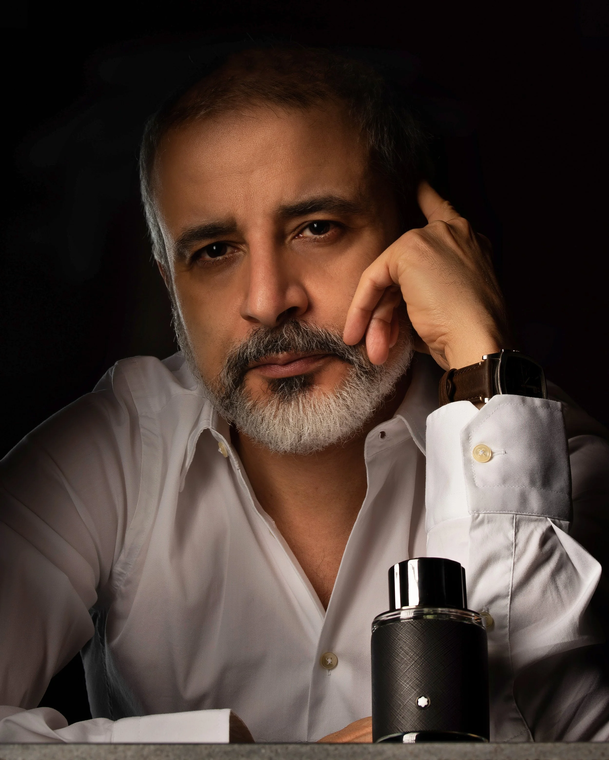 A man with gray hair and beard, wearing a white shirt, is looking directly at the camera with a serious expression. He has a watch on his left wrist and is resting his head on his left hand, with a perfume bottle in front of him.