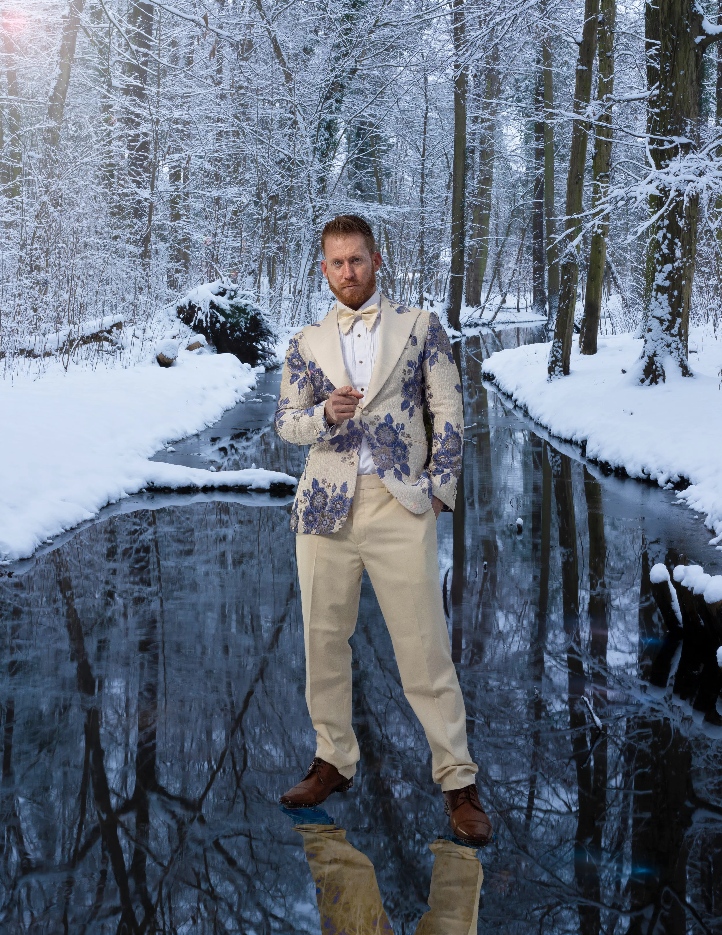 A man wearing a beige suit with blue floral patterns, a white shirt, and a bow tie standing on a frozen, reflective ice surface in a snowy forest with snow-covered trees and a stream in the background.