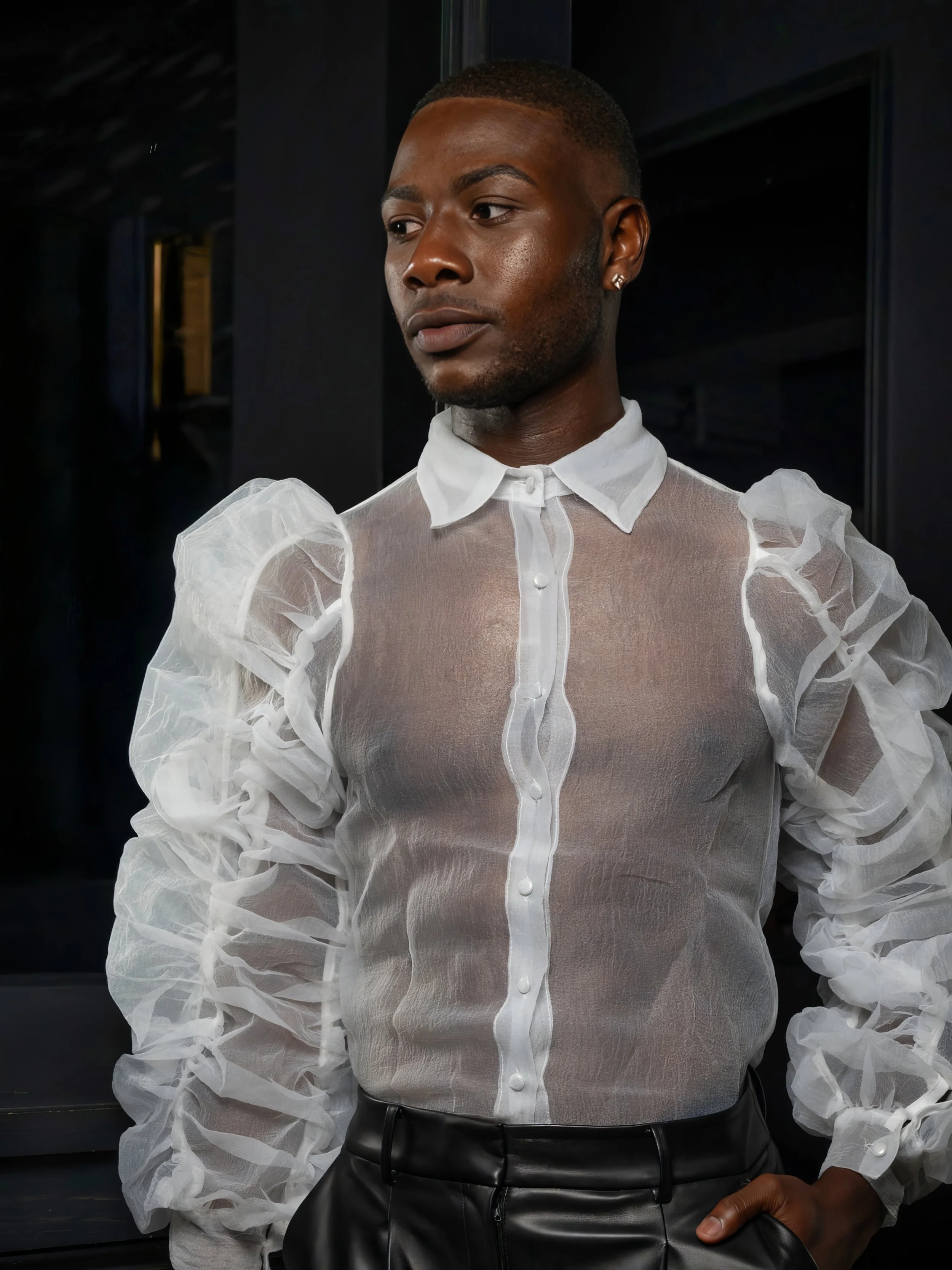 A person with dark skin and short hair wearing a sheer white blouse with puffed sleeves and a high collar, paired with black pants, standing indoors against a dark background.
