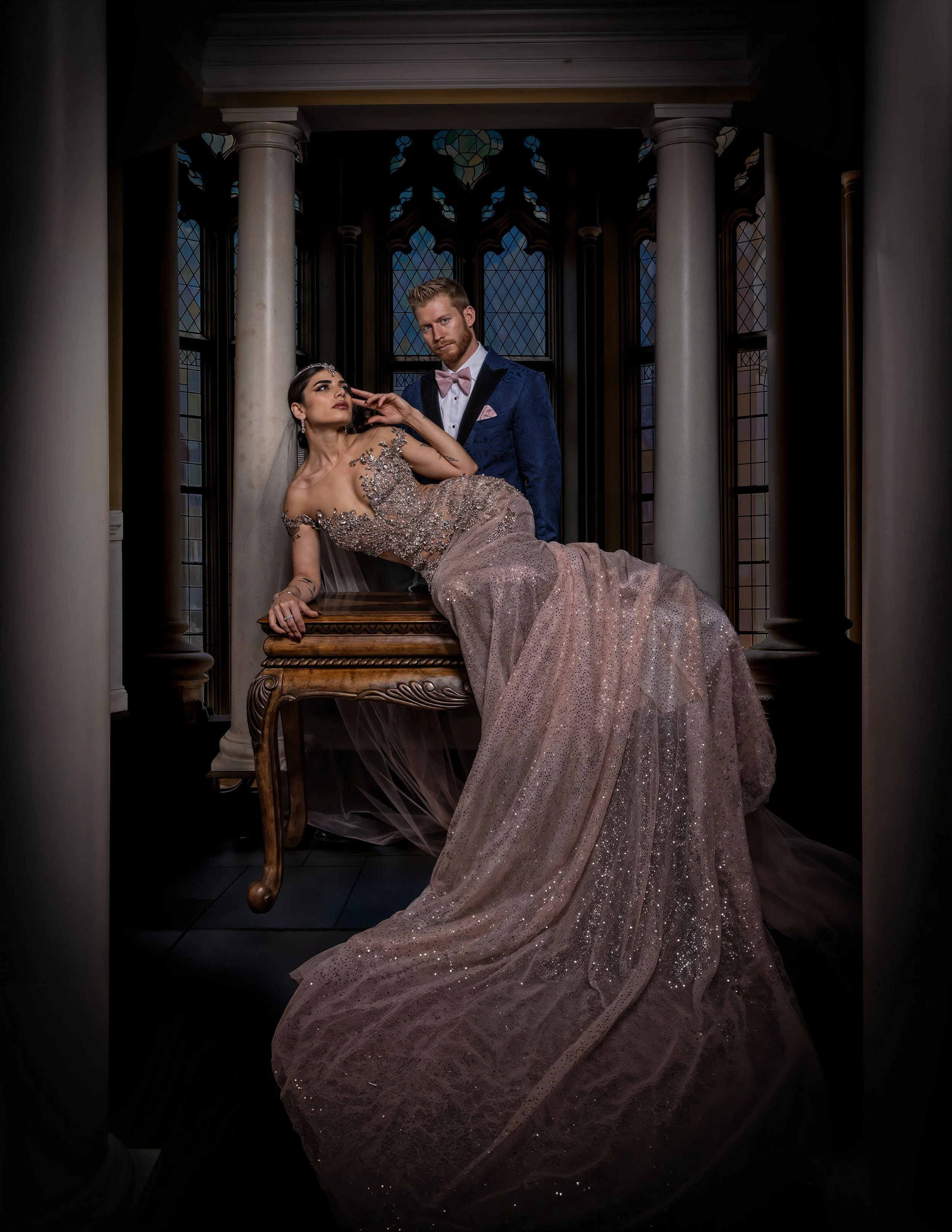 A man and woman in formal attire posing in a gothic-style room with stained glass windows. The woman is sitting on a table, wearing a sparkling gown, and the man stands behind her in a dark blue tuxedo.