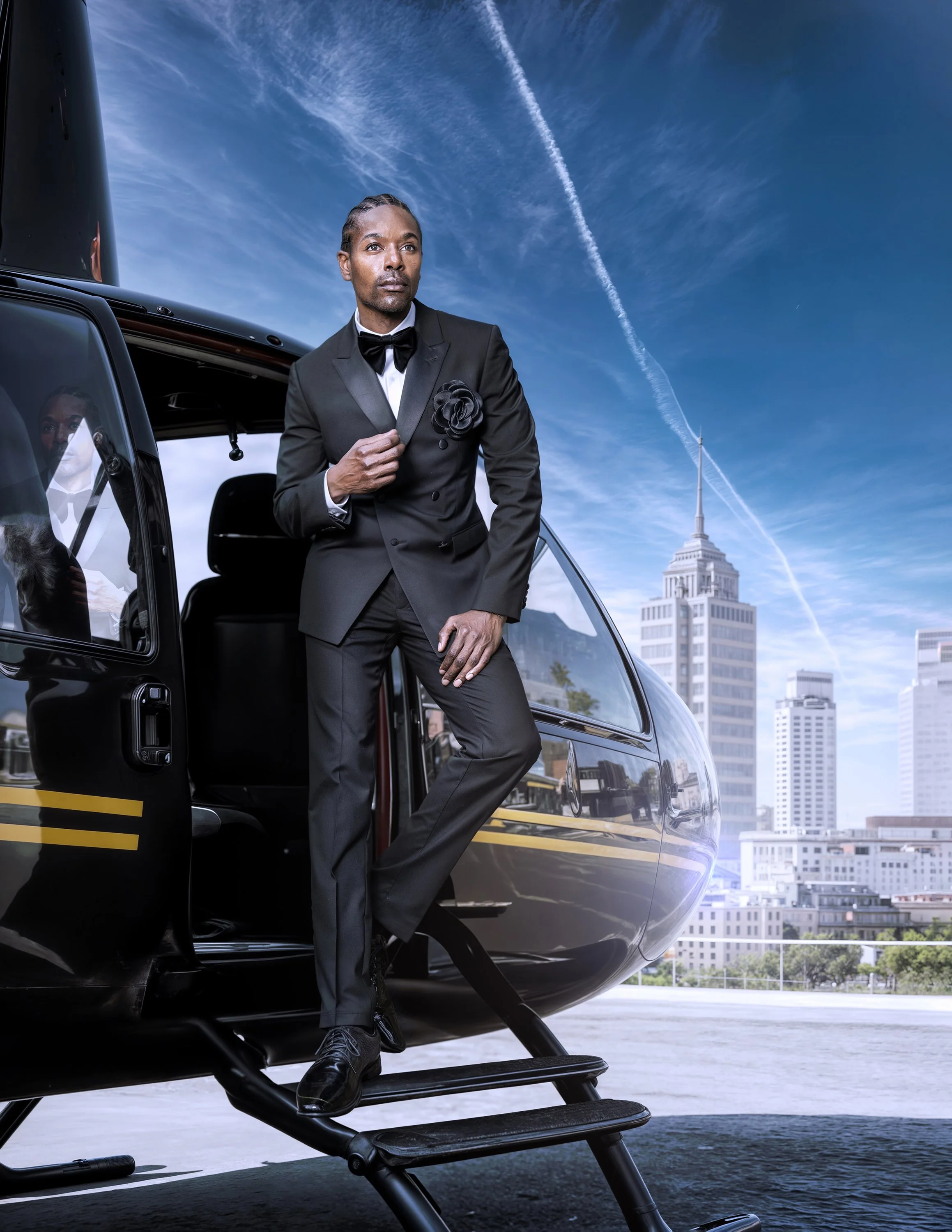 A fashion model in a black tuxedo with a bow tie standing on the steps of a helicopter during the daytime. The city skyline, including the Empire State Building, is visible in the background.