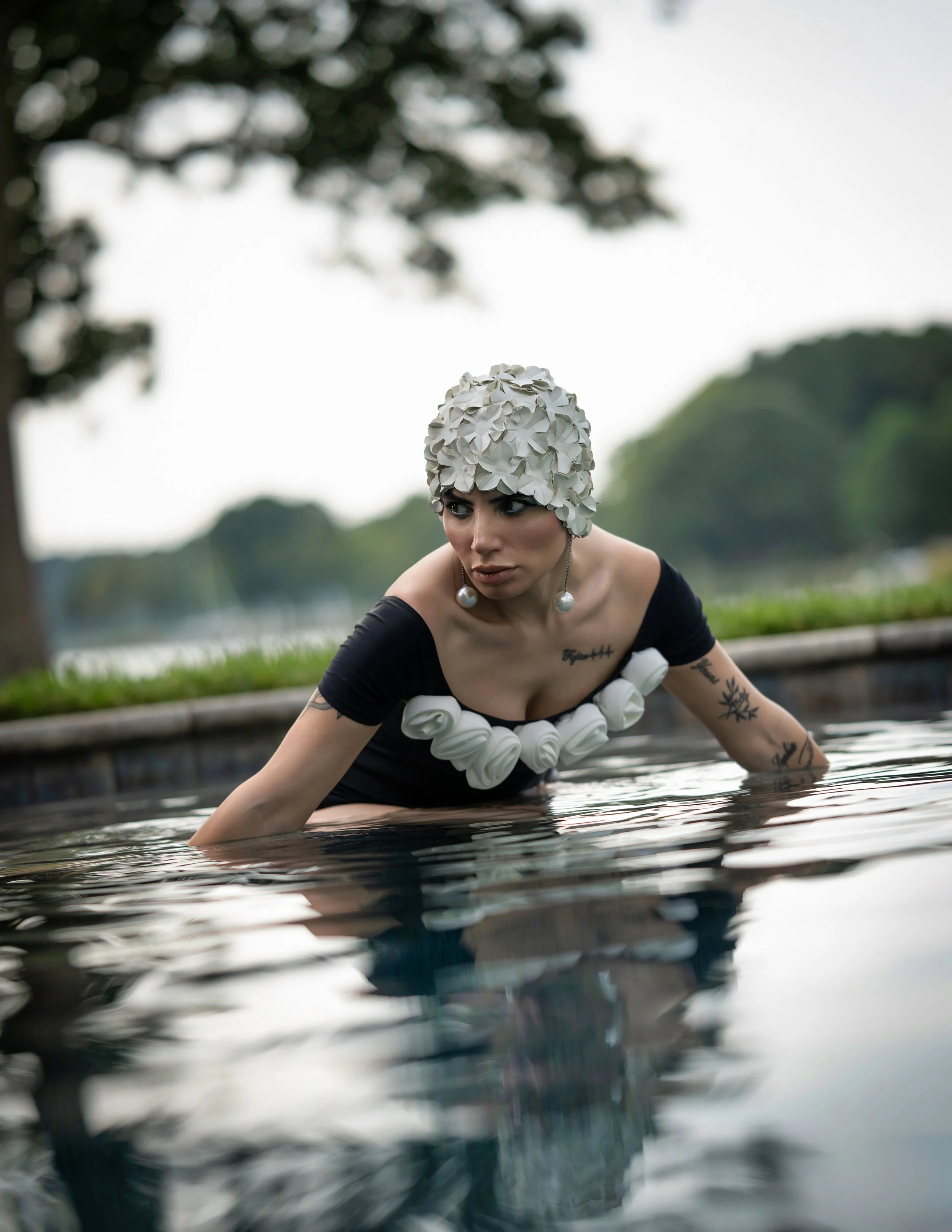 Woman wearing a floral headpiece and black off-shoulder dress with floral embellishments, leaning in a pool with water reaching her chest, outdoors with blurred trees and sky in the background.