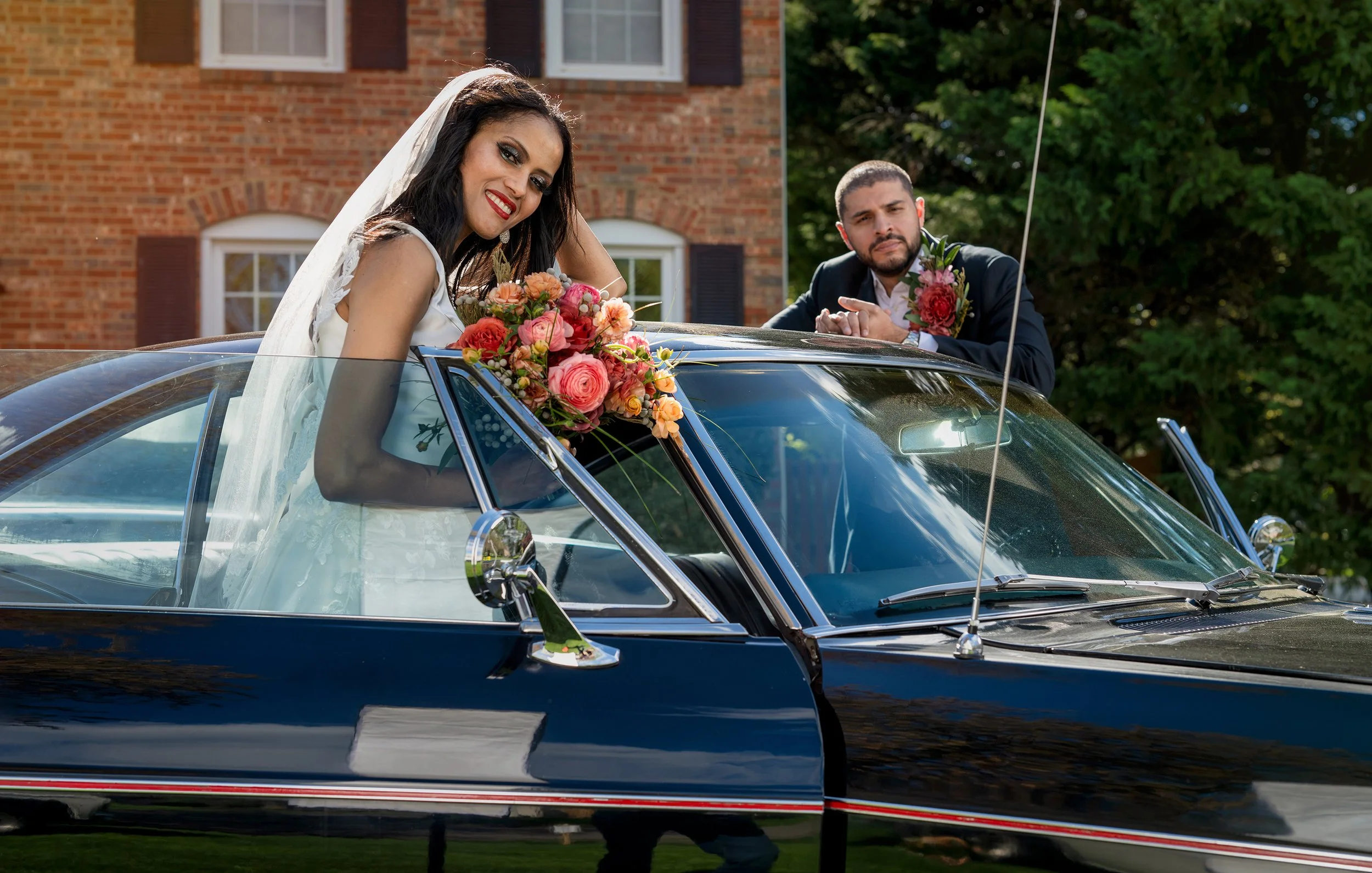 A bride and groom, dressed in wedding attire, sitting in a vintage black car during their wedding celebration outdoors on a sunny day.