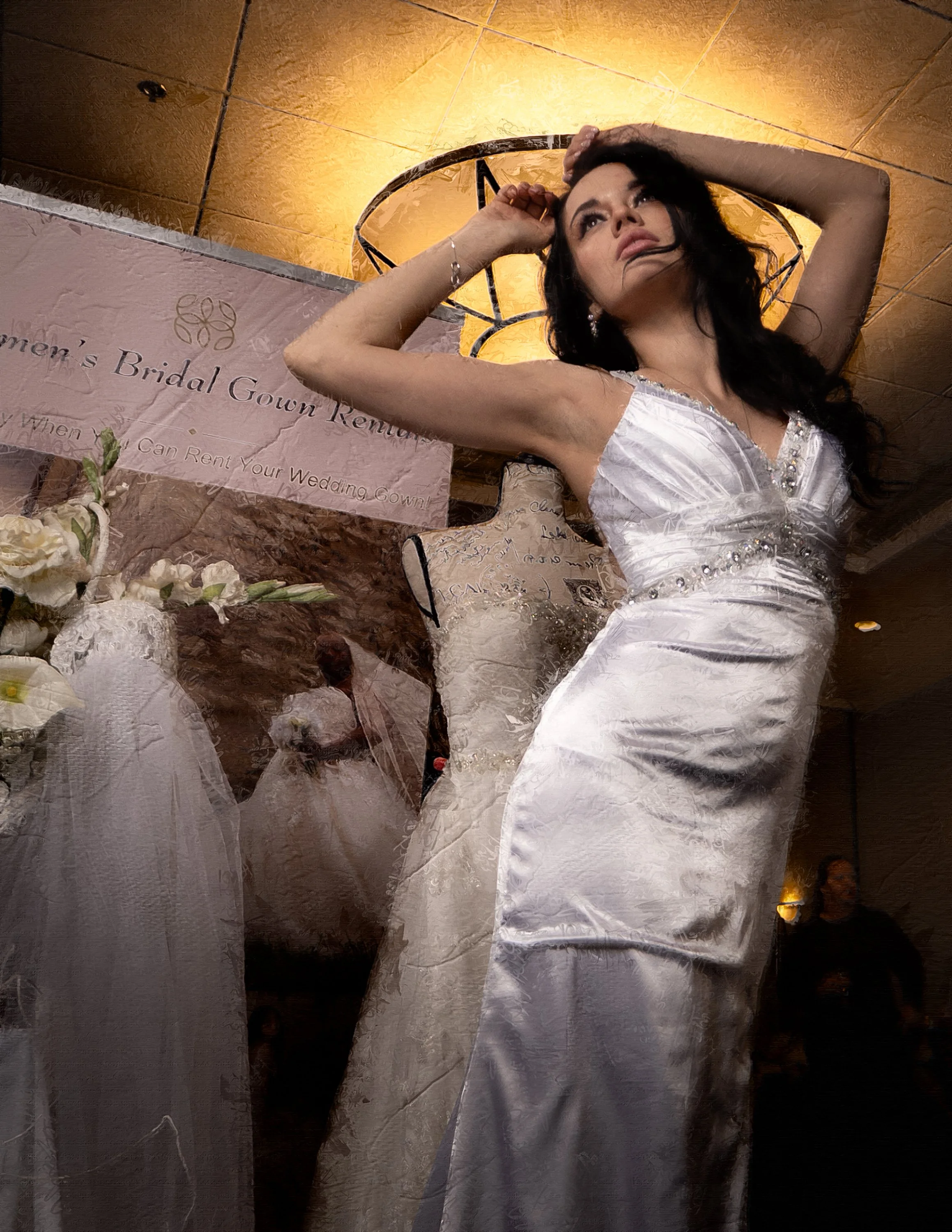 A woman in a white bridal gown posing with her left arm behind her head indoors, with a wedding dress display and a pink sign in the background.