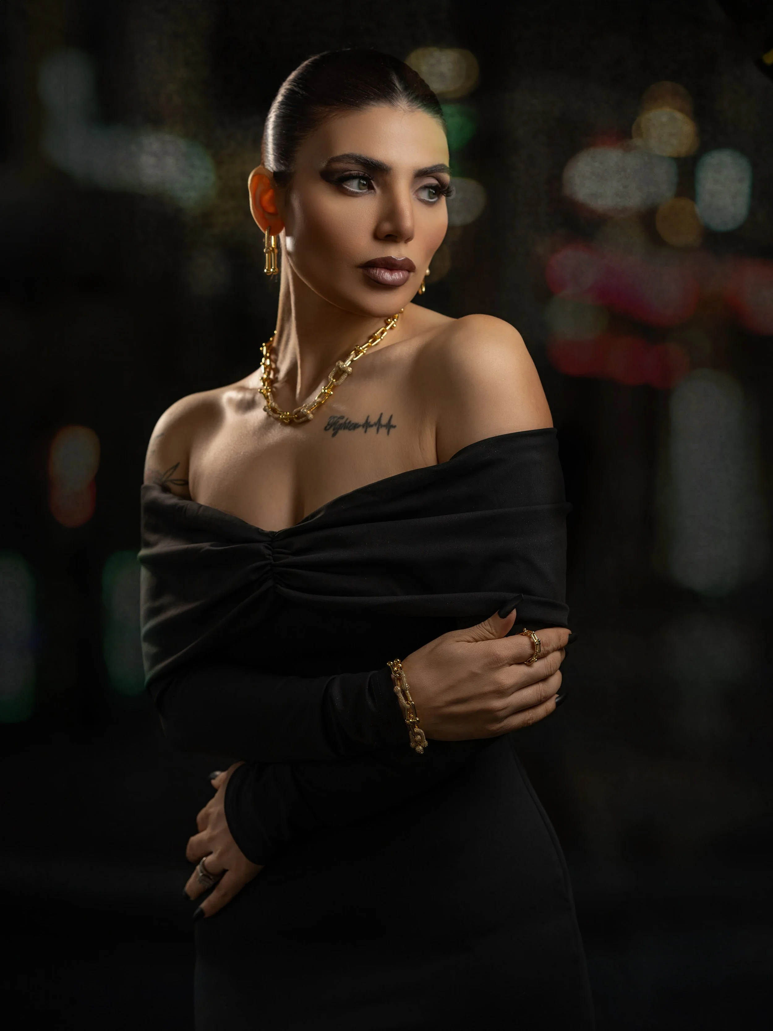 A woman with dark hair styled in a sleek bun, wearing makeup with bold lipstick, adorned with gold jewelry including earrings, a necklace, bracelet, and rings, dressed in an off-shoulder black dress, with a cityscape blurred in the background at nigh
