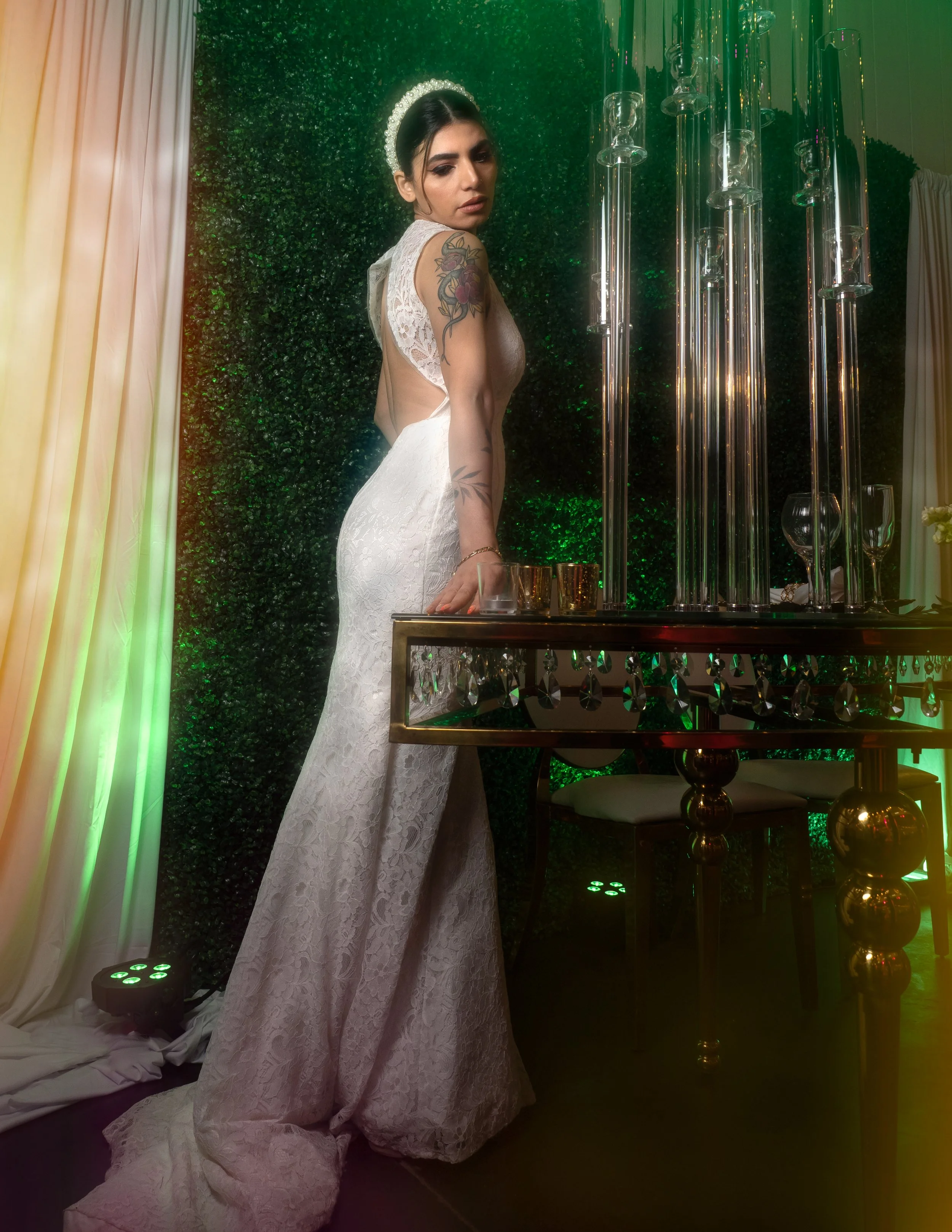 A woman in a white lace dress with open back, wearing a headband, stands next to a table with candles and glasses, in front of a green leafy backdrop, at a formal event.
