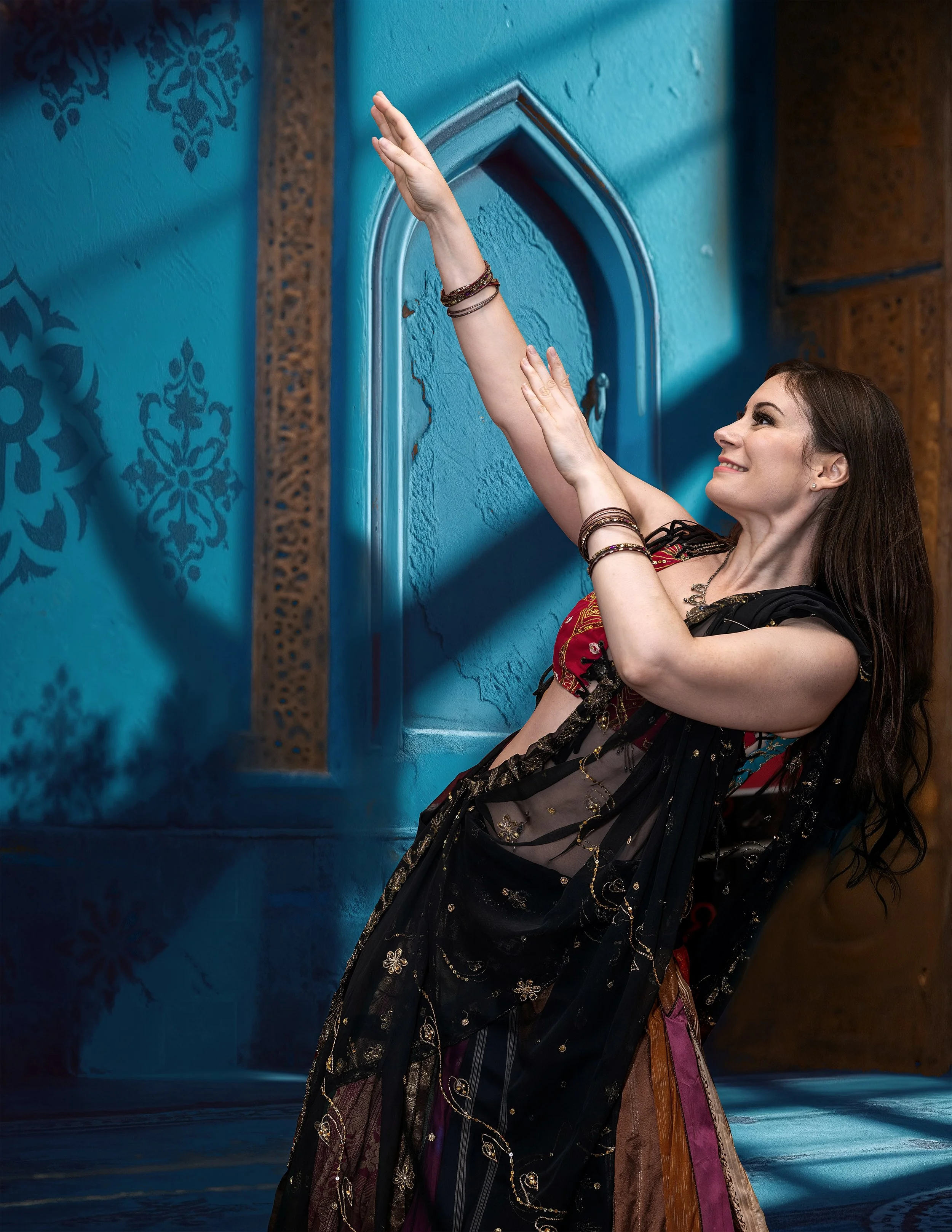 Woman in colorful traditional dress posing with their arm raised in front of a blue wall with ornate designs.