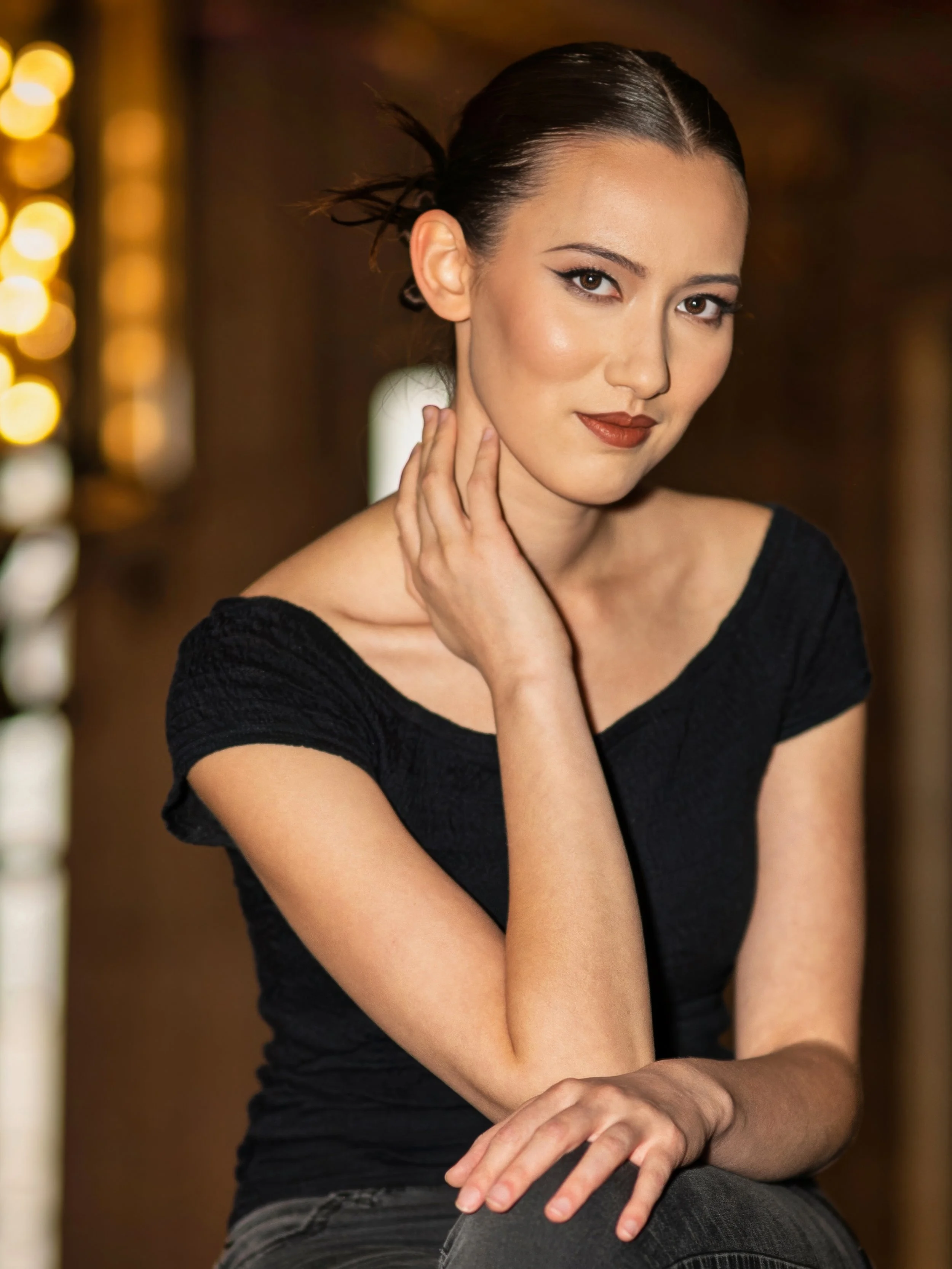 A woman with dark hair and makeup, wearing a black top, sitting with her hand resting on her neck and her other hand on her knee, in a warmly lit indoor setting.