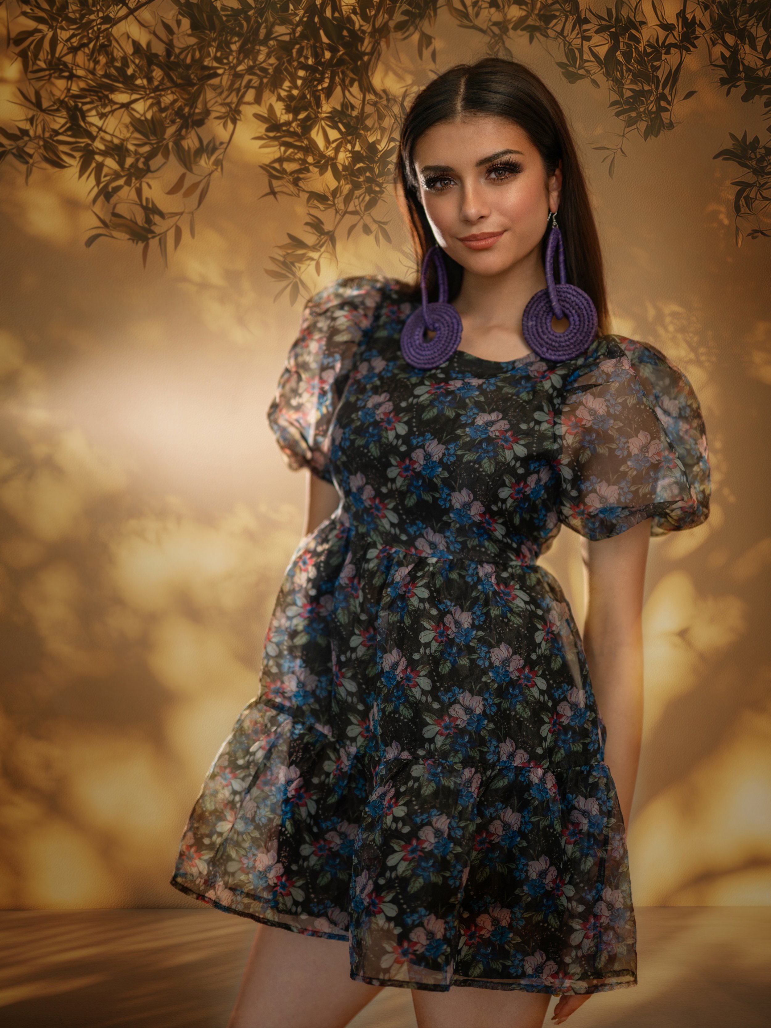 A woman with long dark hair wearing large purple circular earrings and a black floral dress with puffed short sleeves, standing in front of a warm, abstract nature background with tree branches and blurred leaves.