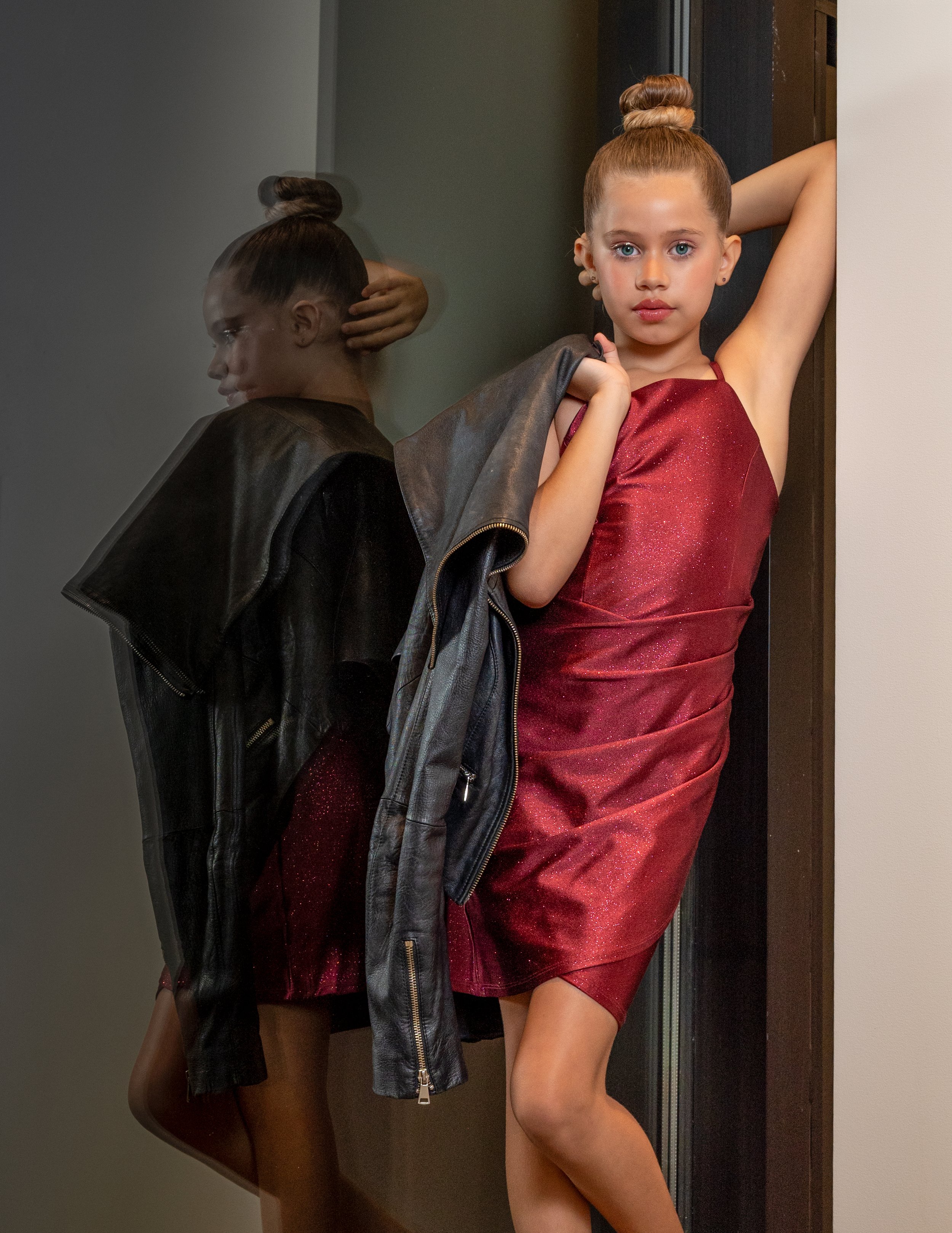 A young woman in a red dress with her hair in a bun is posing with one arm raised, holding a black leather jacket over her shoulder, standing beside a mirror that reflects her image.