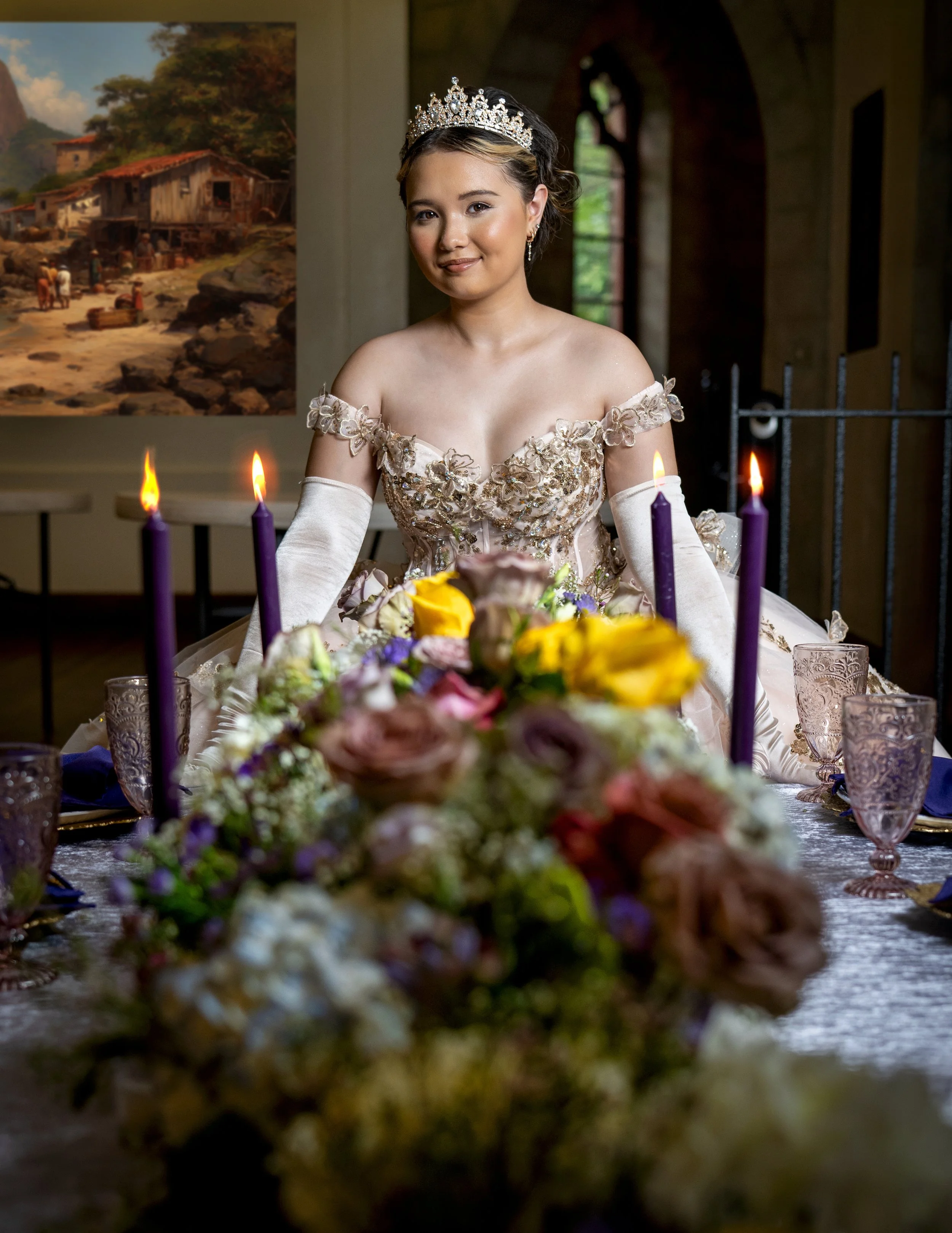 Young woman in an elegant gown and tiara sitting at a decorated table with flowers and candles, indoors with a large painting in the background.
