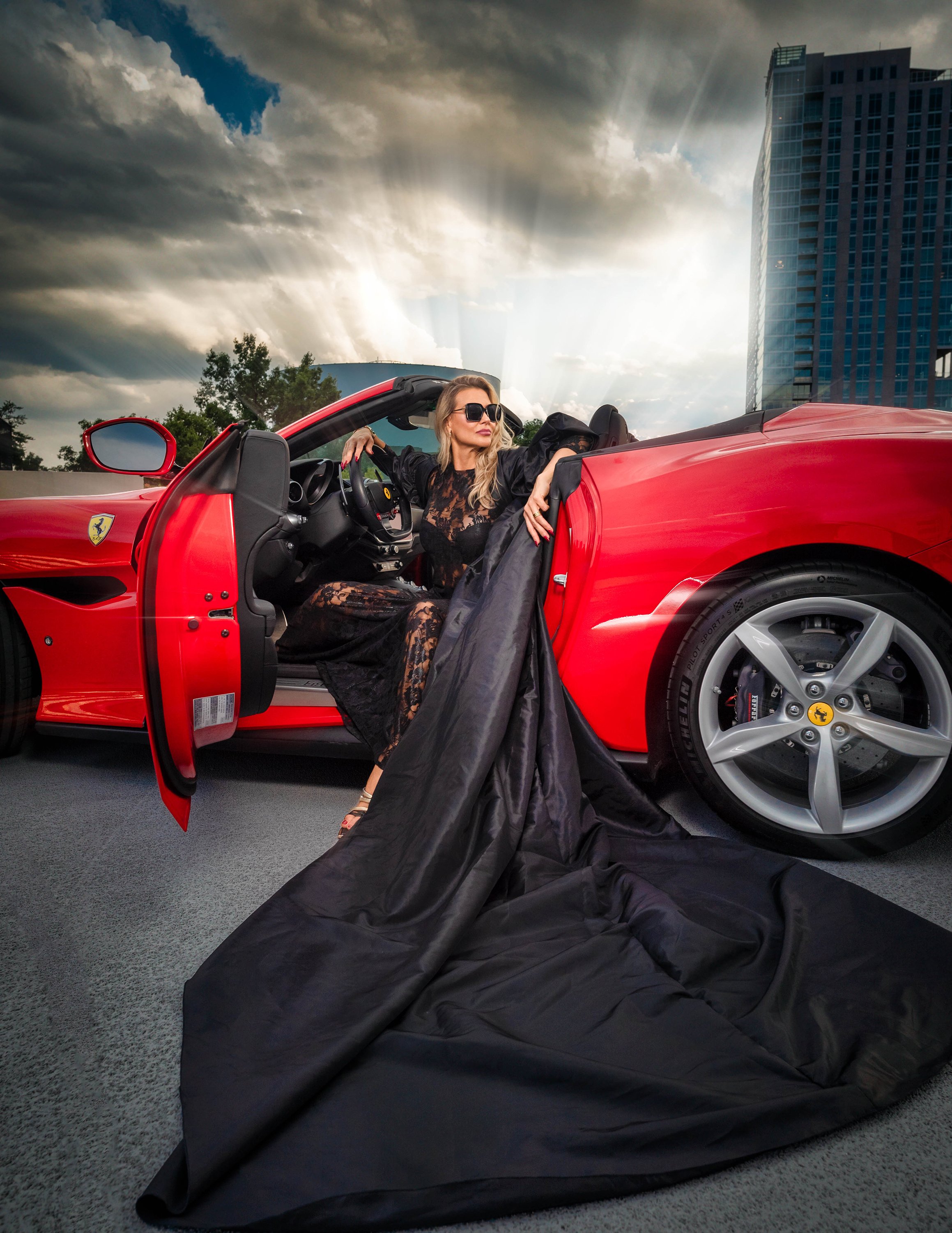A fashion model in sunglasses and a black lace dress sitting in a red Ferrari with the door open, on a rooftop parking lot with a dark sky and tall buildings in the background.