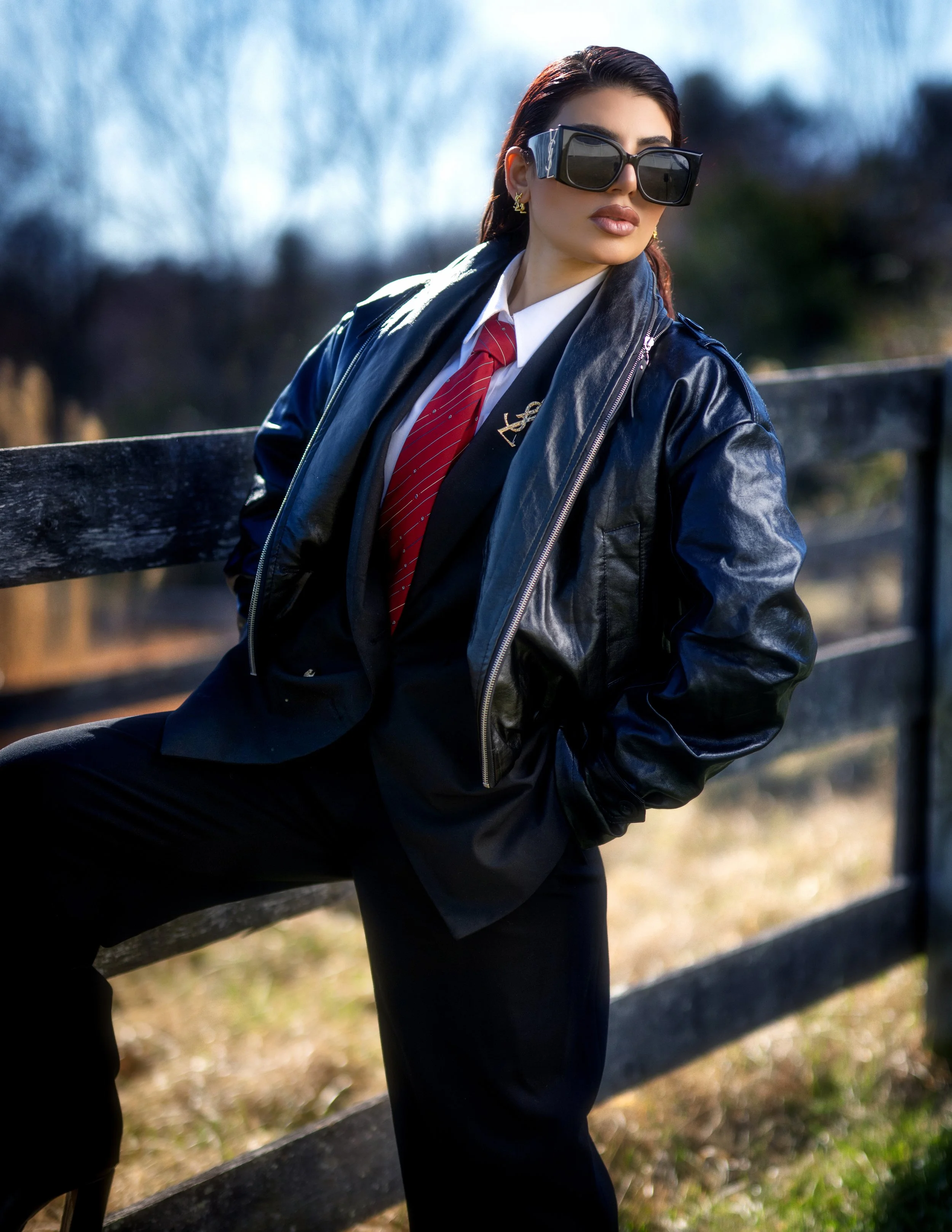 A woman wearing large black sunglasses, a black leather jacket, a white shirt, a red tie, and black pants standing outdoors near a wooden fence.