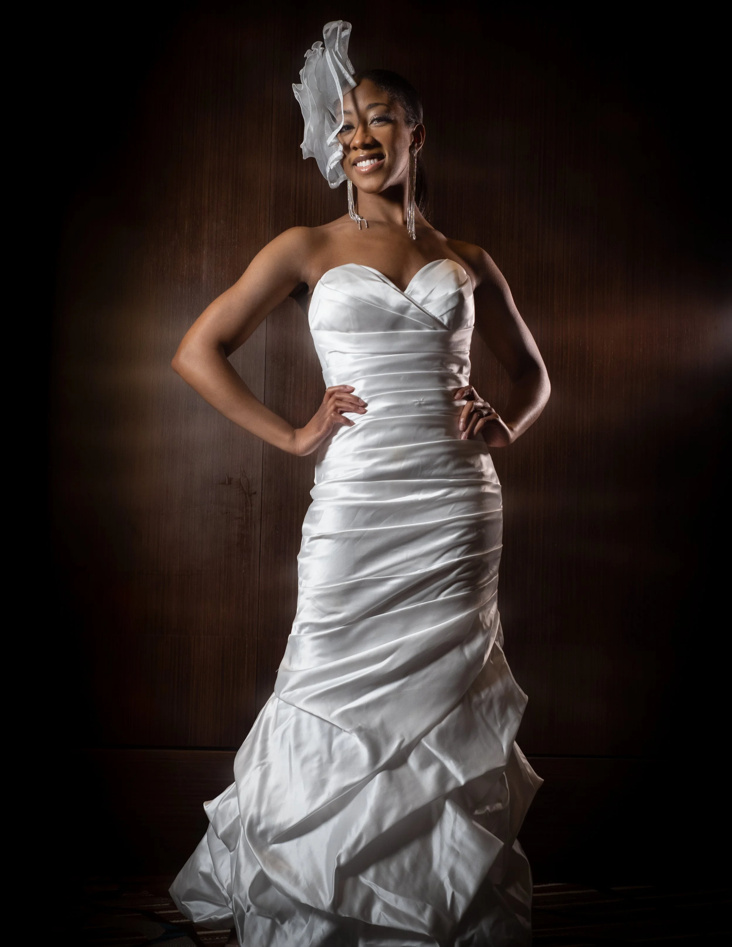 A woman in a white wedding dress with a strapless sweetheart neckline, standing with her hands on her hips, smiling confidently, wearing a large white headpiece and long earrings, against a dark wood-paneled wall.