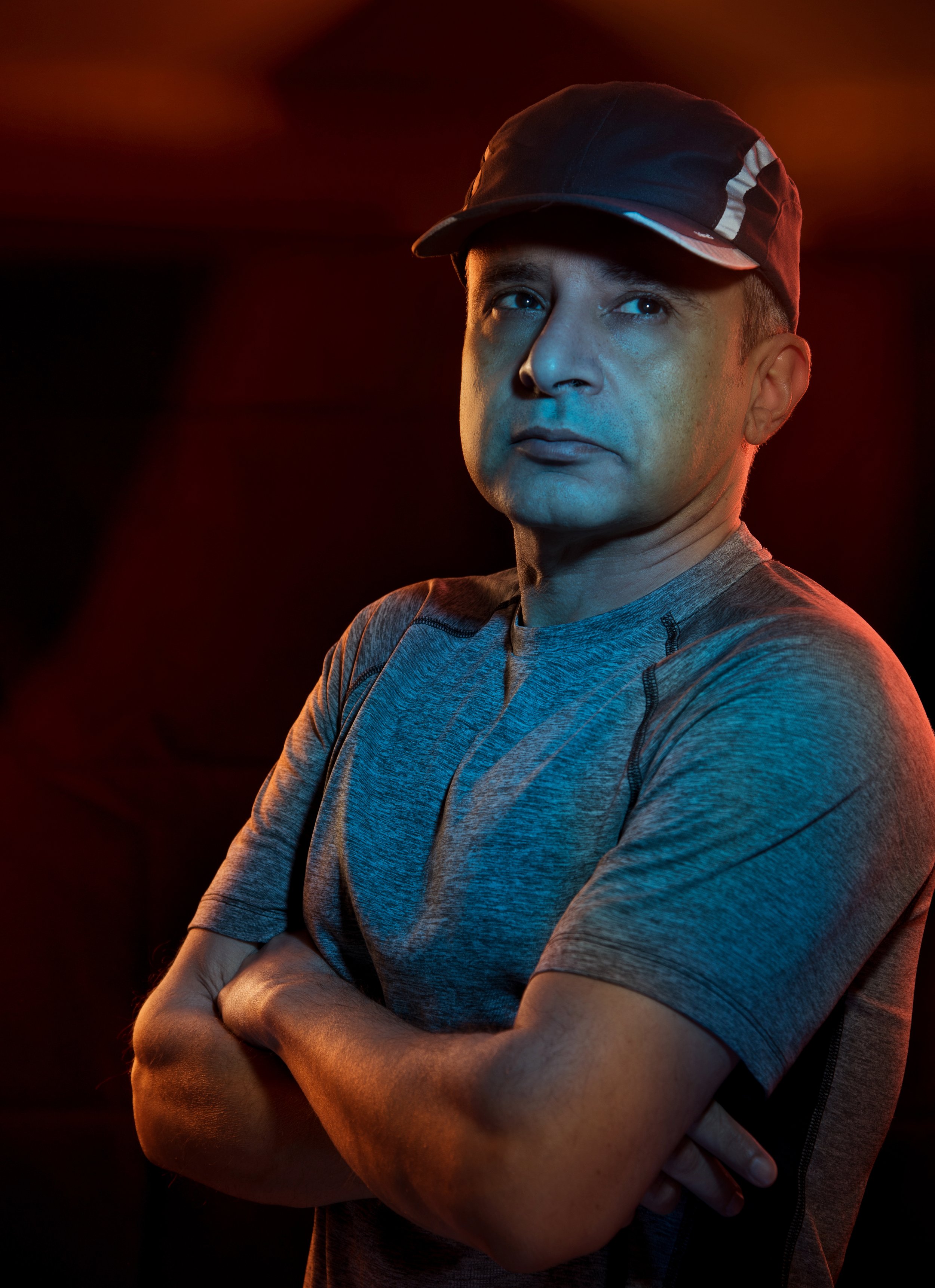 A man wearing a cap and athletic shirt standing with arms crossed in a dark setting with red and blue lighting.