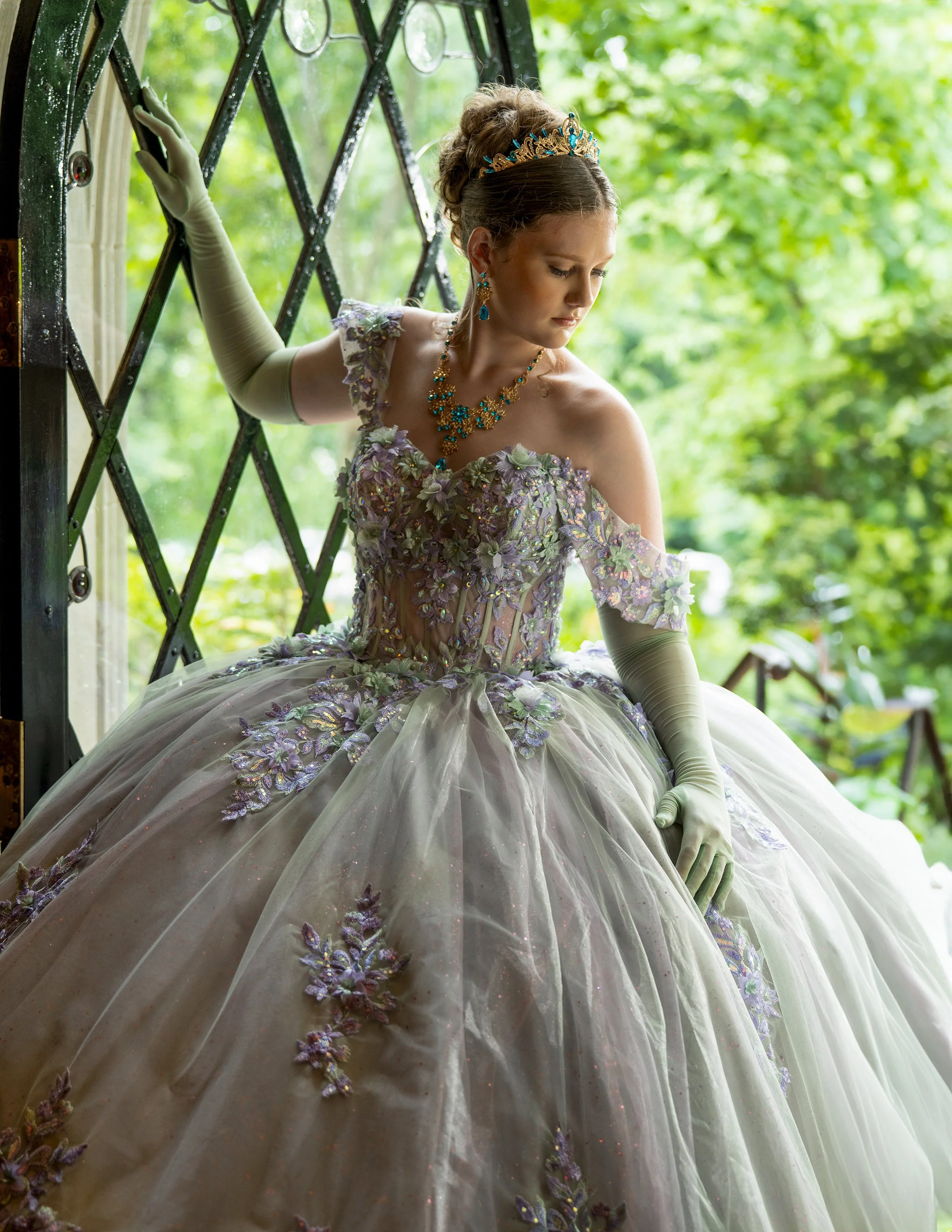 A woman dressed in an elaborate, lavender and cream-colored ballgown with floral embroidery and embroidery details, wearing jewelry and a tiara, standing near a window with lush green foliage outside, looking down thoughtfully.