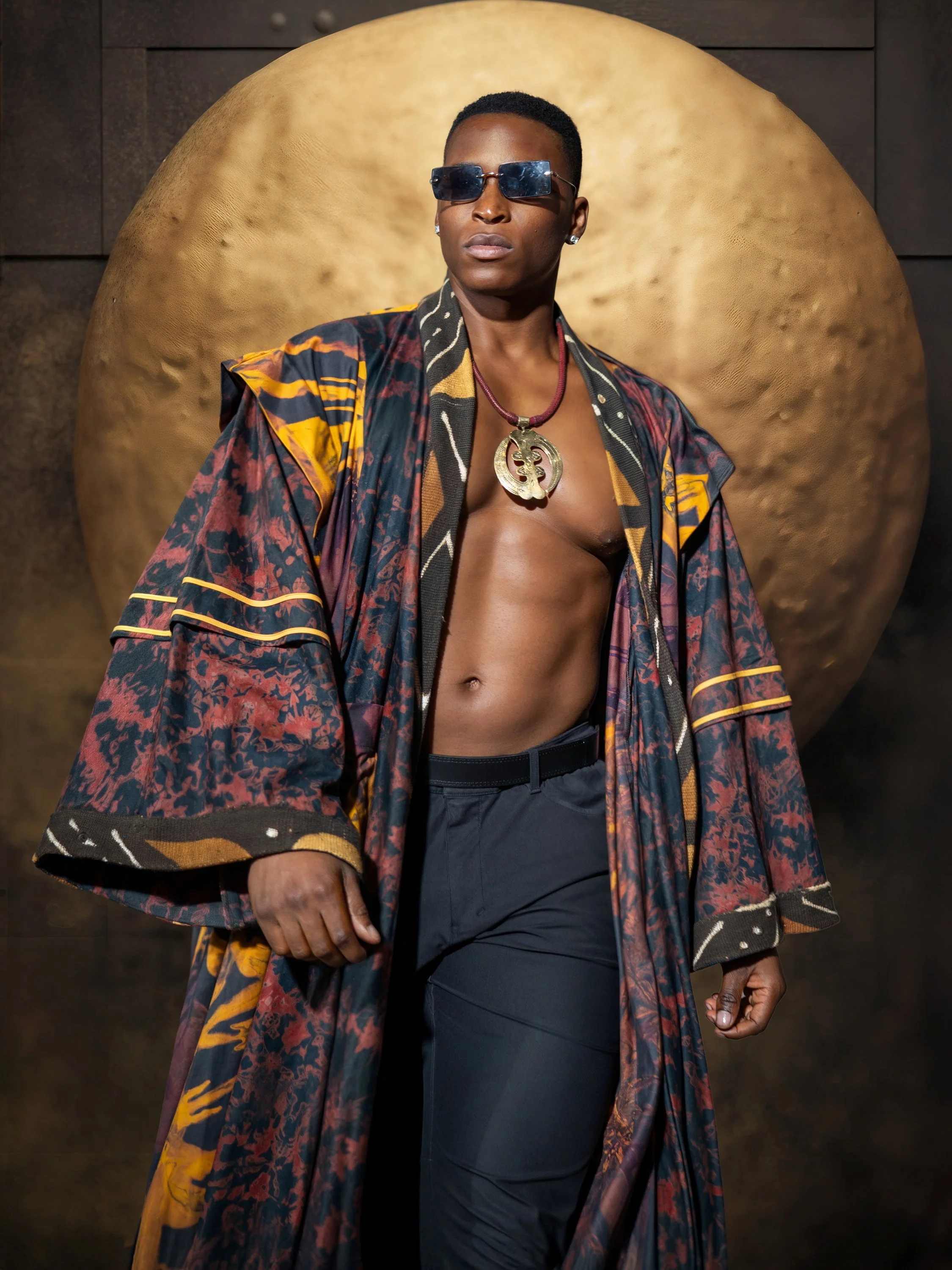 Individual wearing sunglasses, a large colorful patterned coat, and a large necklace, standing in front of a dark background with a large gold sphere.