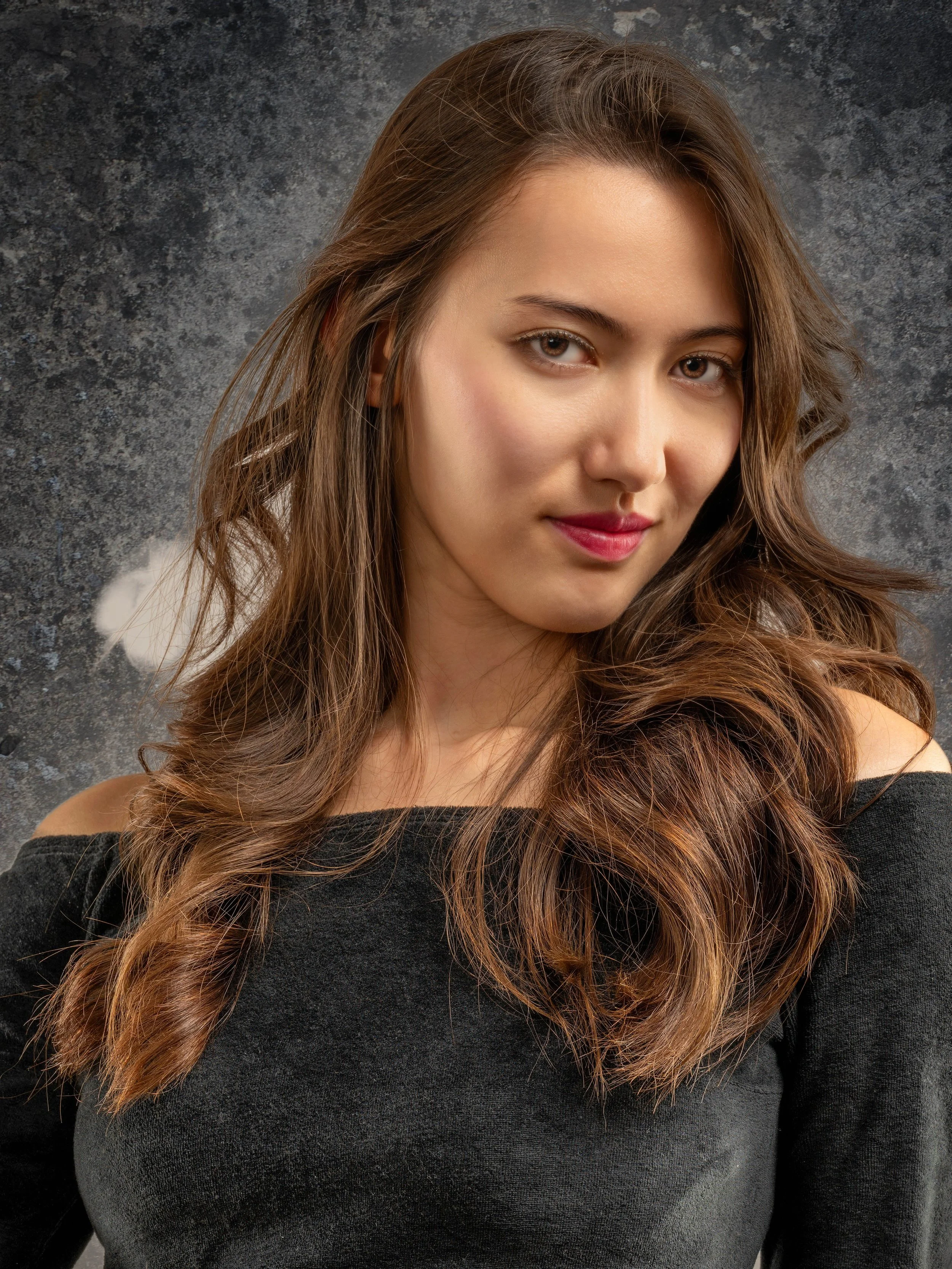 A woman with light skin and long, wavy brown hair wearing a black off-shoulder top, looking at the camera with a slight smile, dark textured background.