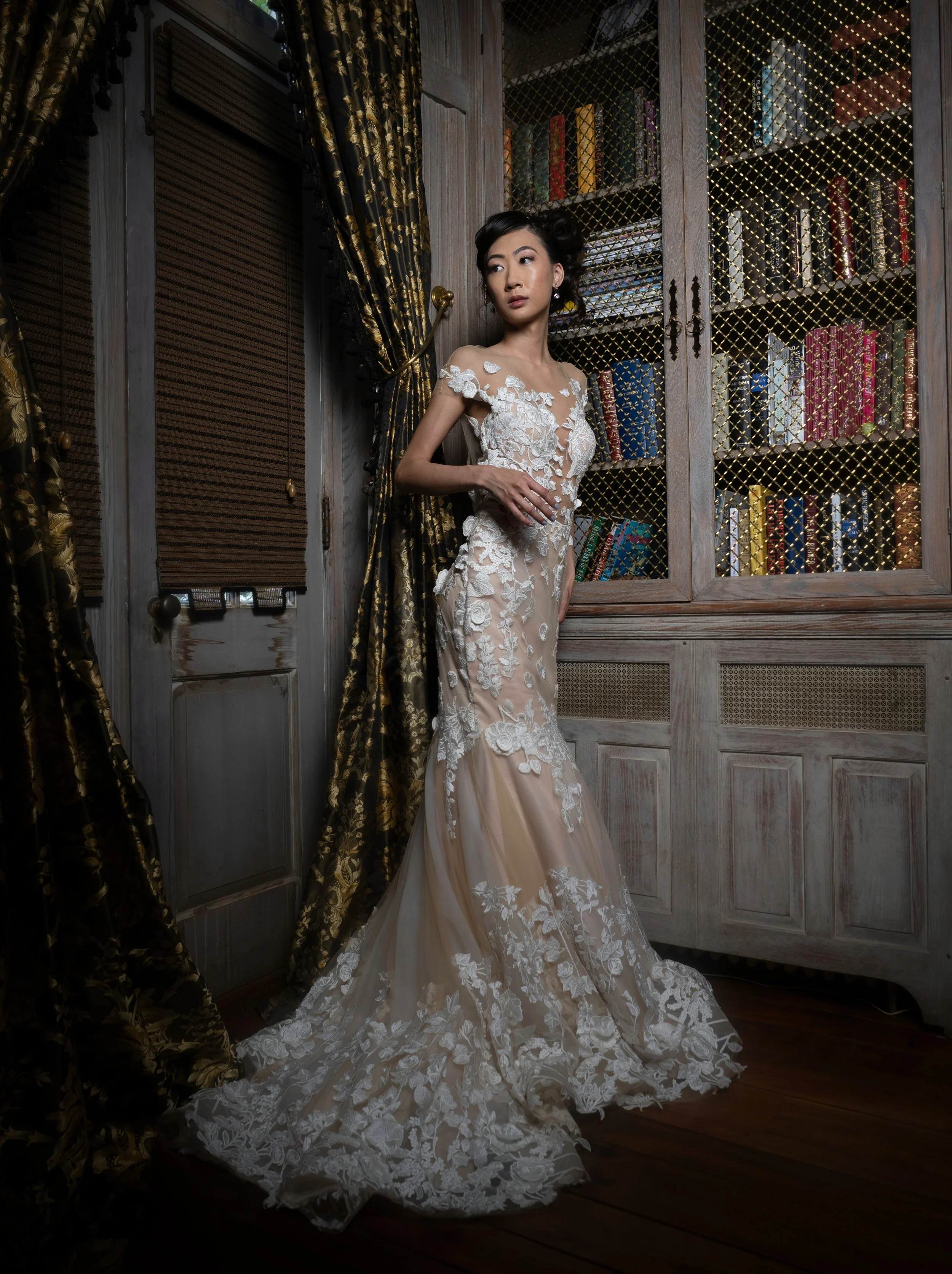 A woman in an elegant, sheer lace wedding gown with floral embroidery, standing in a vintage-style library or study with dark wood furniture, gold curtain rods, and a glass-front bookshelf filled with books.