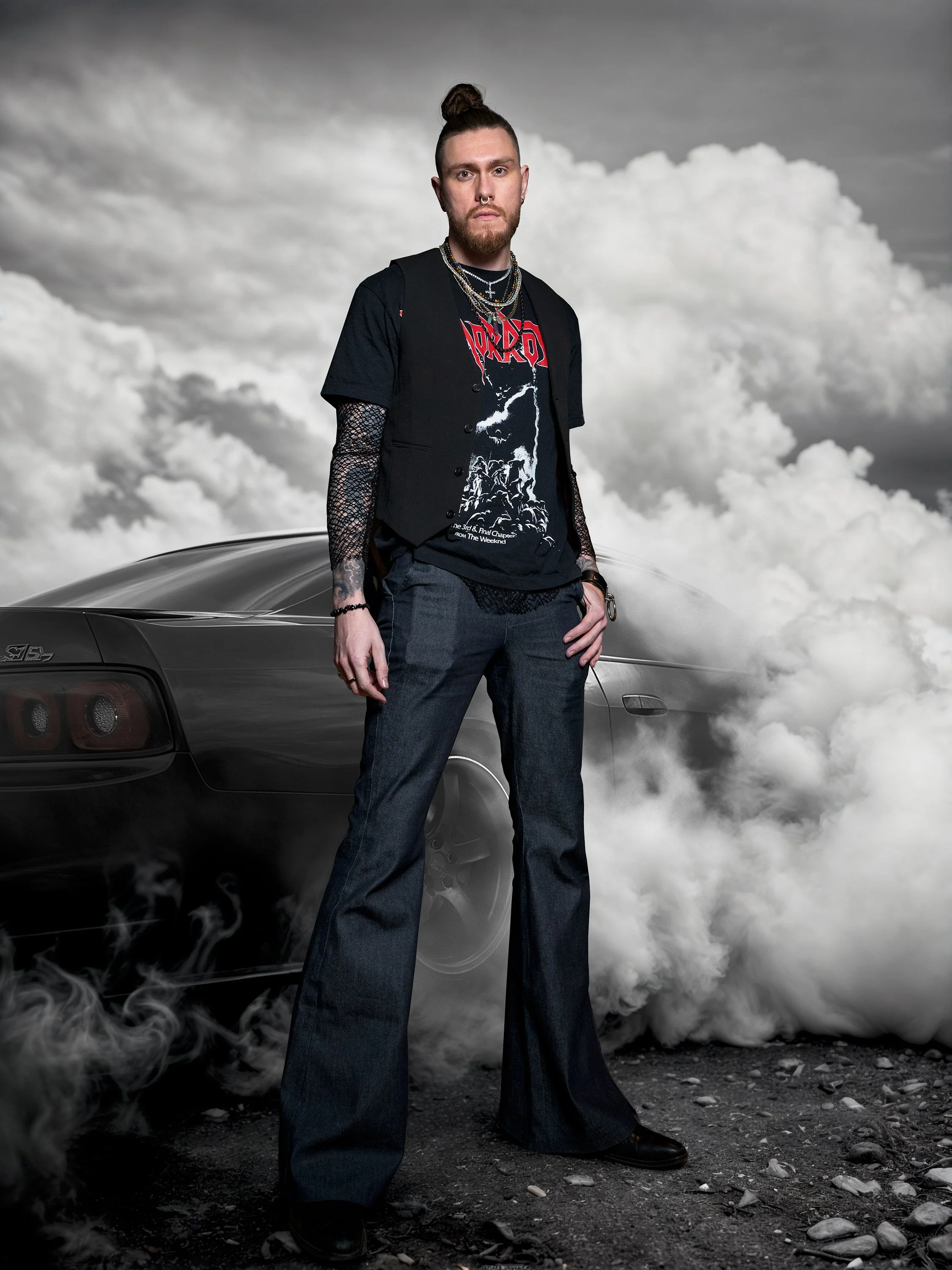 A man with a beard and a top knot hairstyle standing in front of a black sports car with smoke around him, under a cloudy sky. He wears a black t-shirt with a graphic design, a black vest, dark jeans, and jewelry.