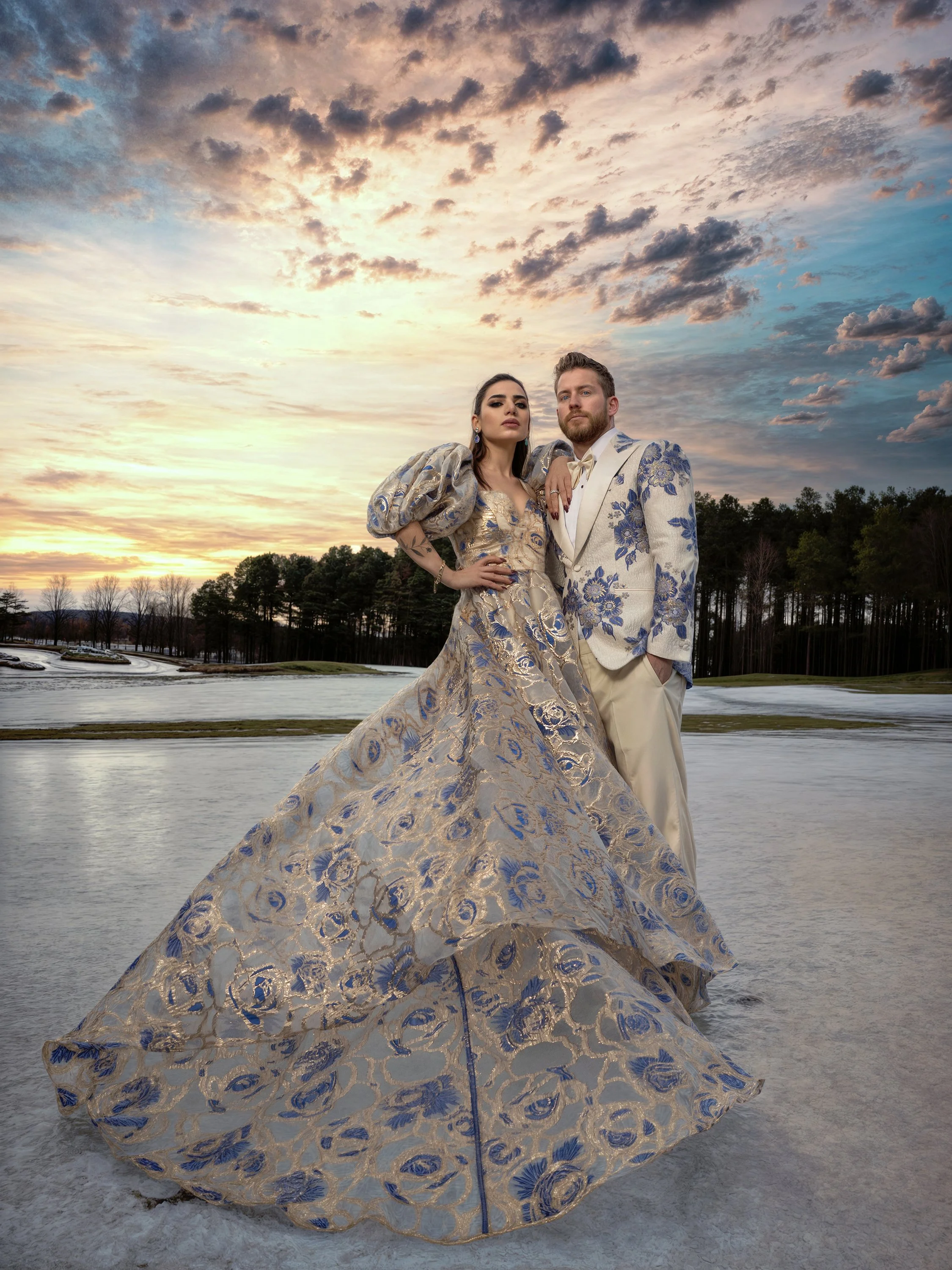 A woman and a man dressed in elaborate formal attire standing outdoors on a winter landscape at sunset.
