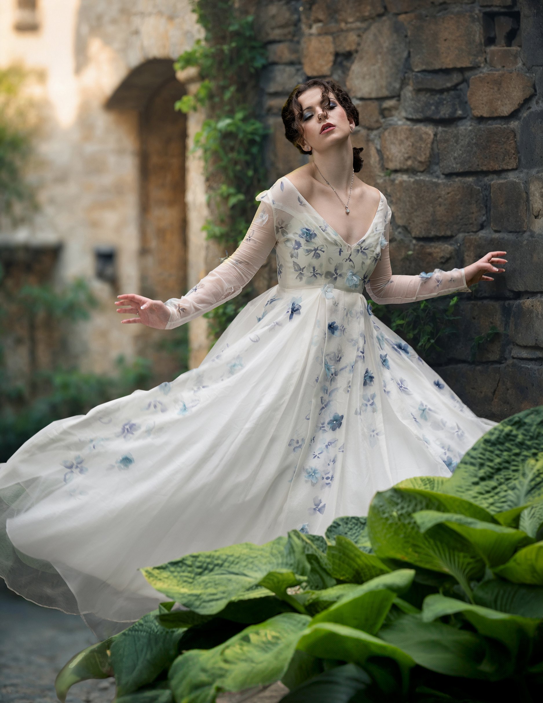 A woman in a vintage white dress with blue flower embroidery poses outdoors against a stone wall, surrounded by greenery.
