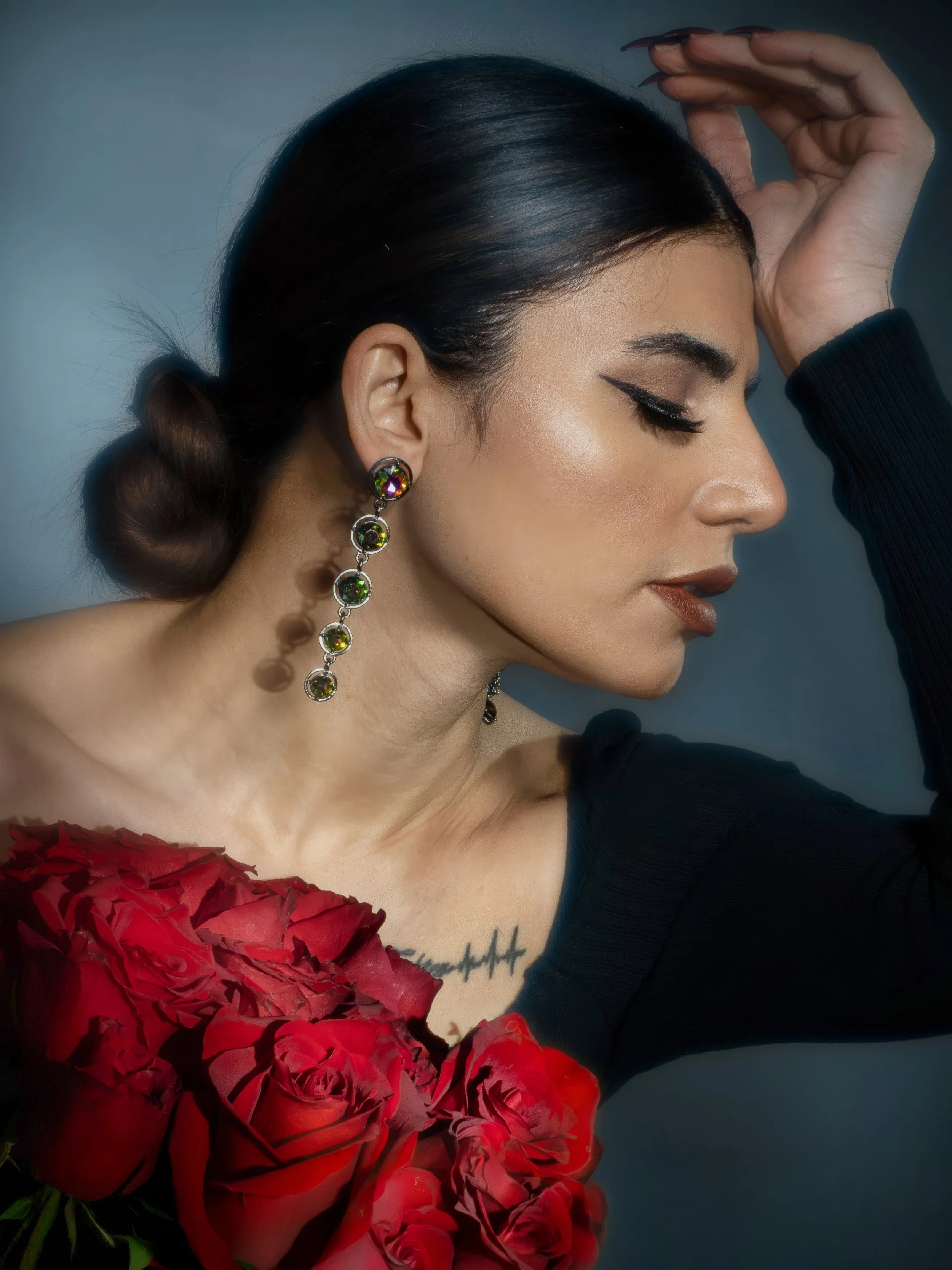 A woman with dark hair styled in a bun, wearing long, multicolored gemstone earrings, matte brown lipstick, thick eyeliner, and a tattoo on her collarbone, is holding her head with her left hand. In front of her, there is a bouquet of red roses.