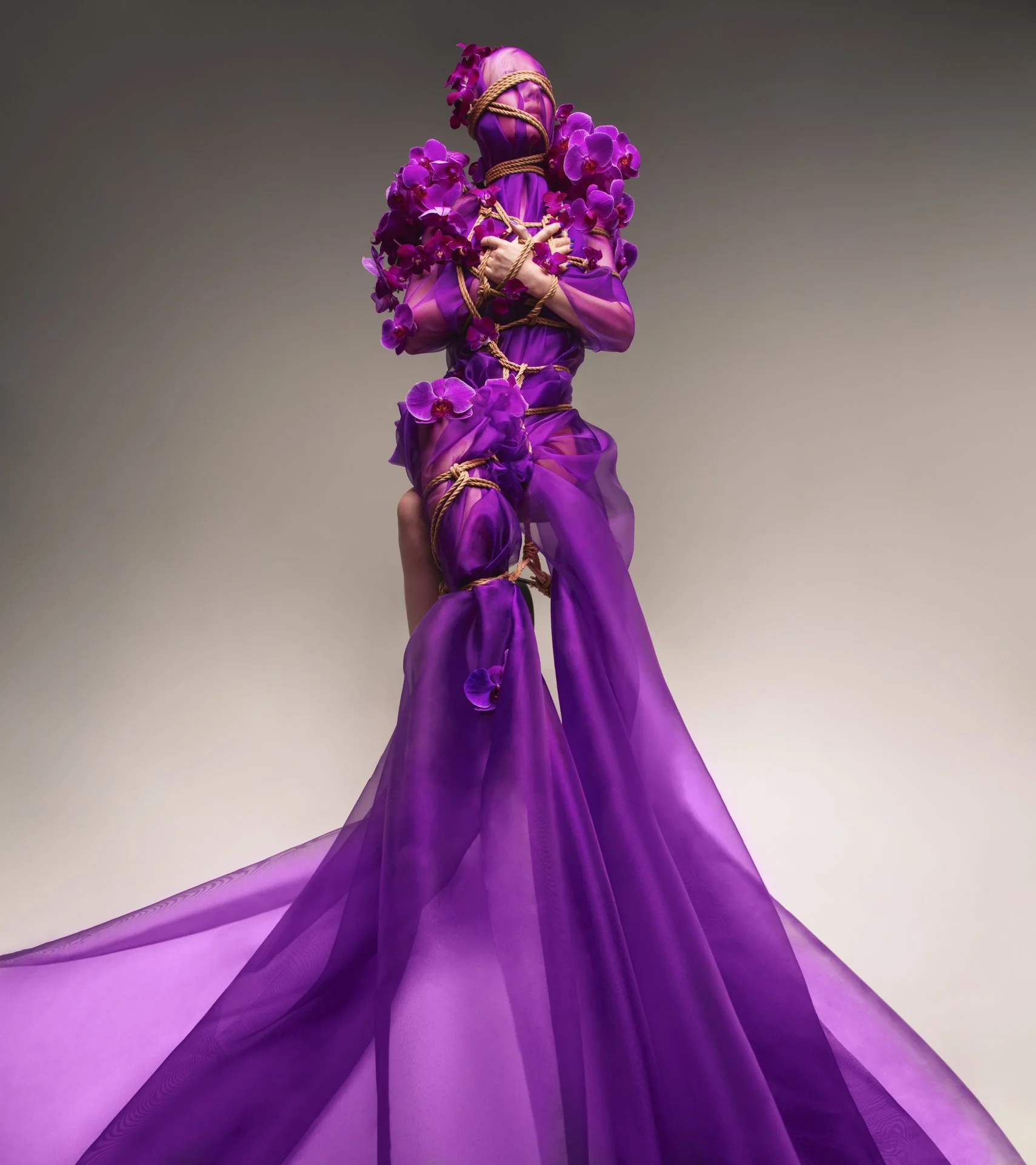 Fashion model in an elaborate purple gown embellished with orchids and tied with rope, against a plain gradient background.