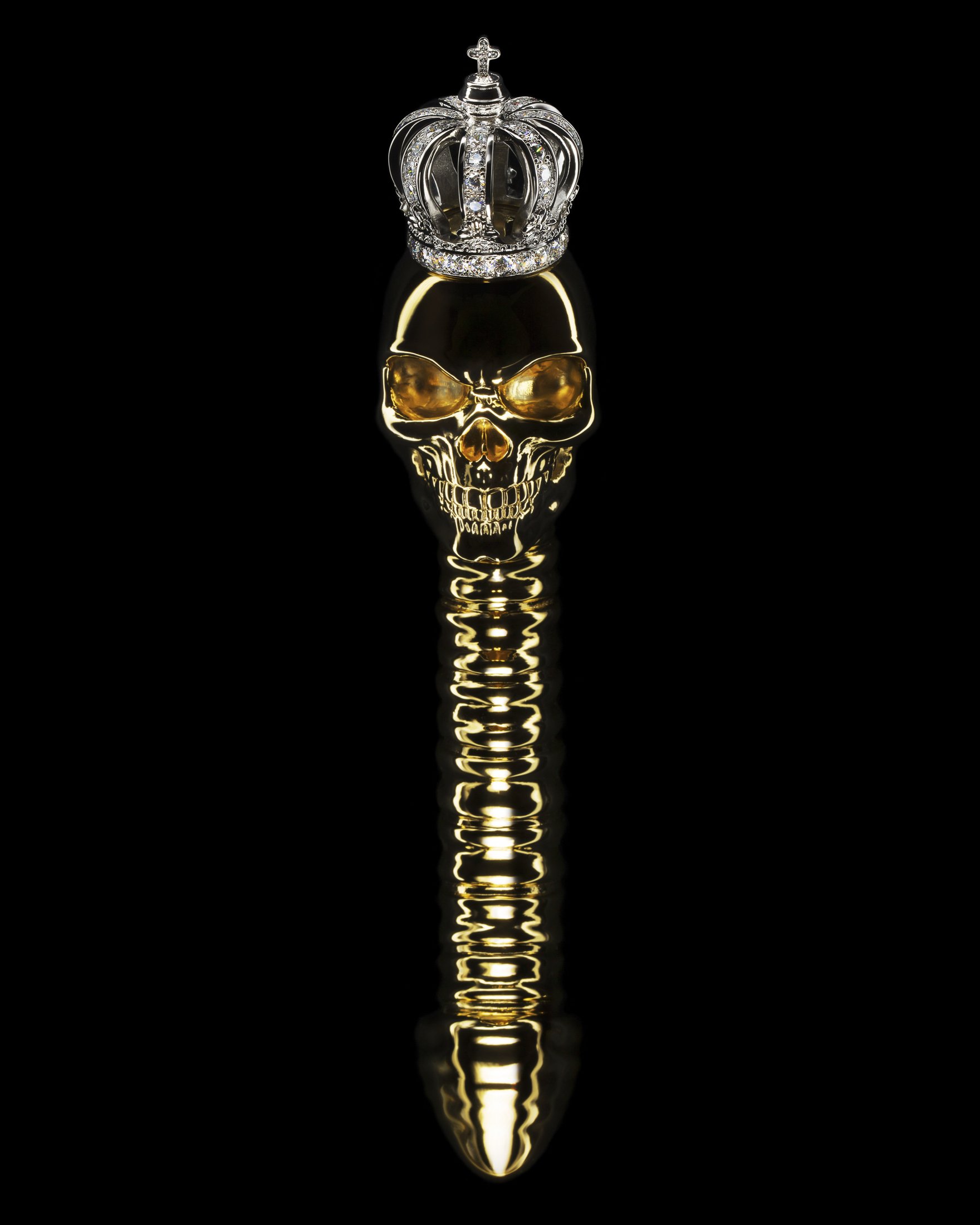Gold skull-shaped object with a diamond-encrusted crown on top, reflected on a glossy surface, against a black background.