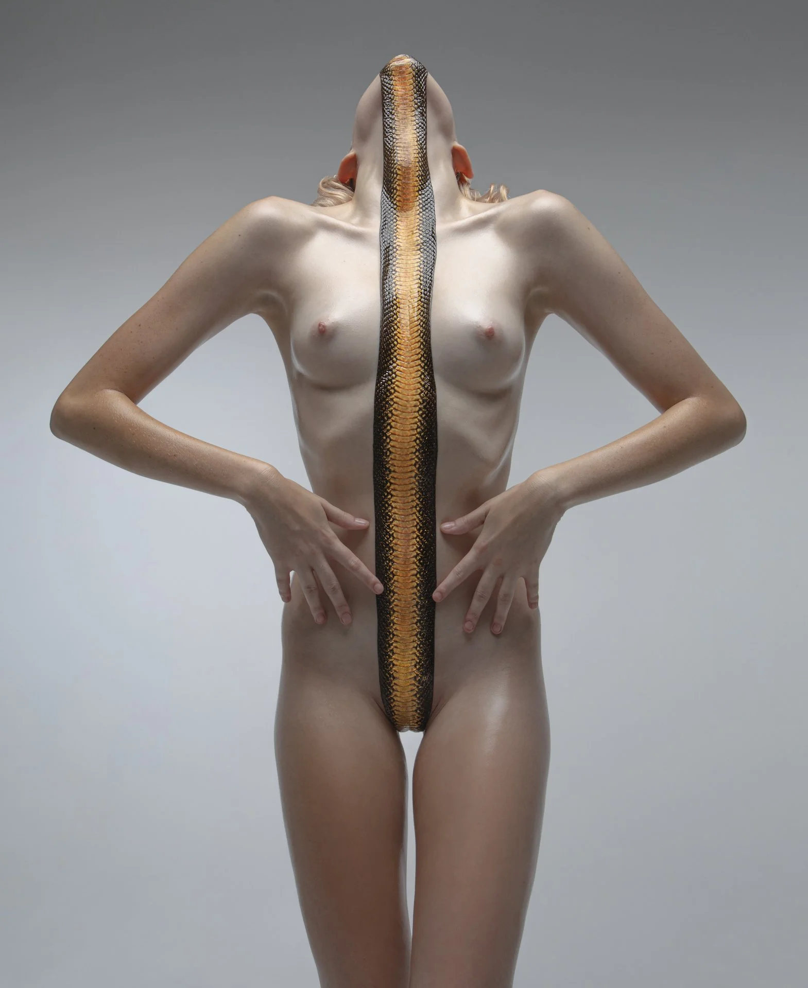 A nude woman with a snake running down her face and chest, covering her face, with her hands resting on her hips.