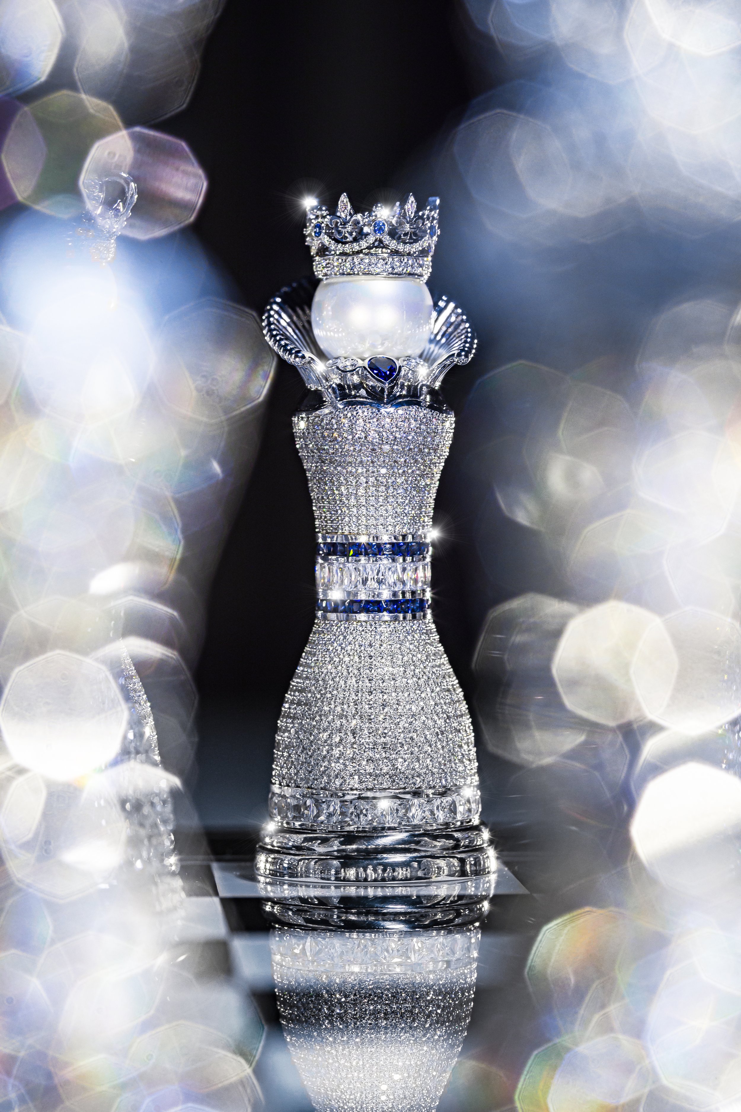 A luxurious chess piece designed like a queen, adorned with diamonds, with a crown on top, set on a reflective surface with colorful bokeh lights surrounding it.