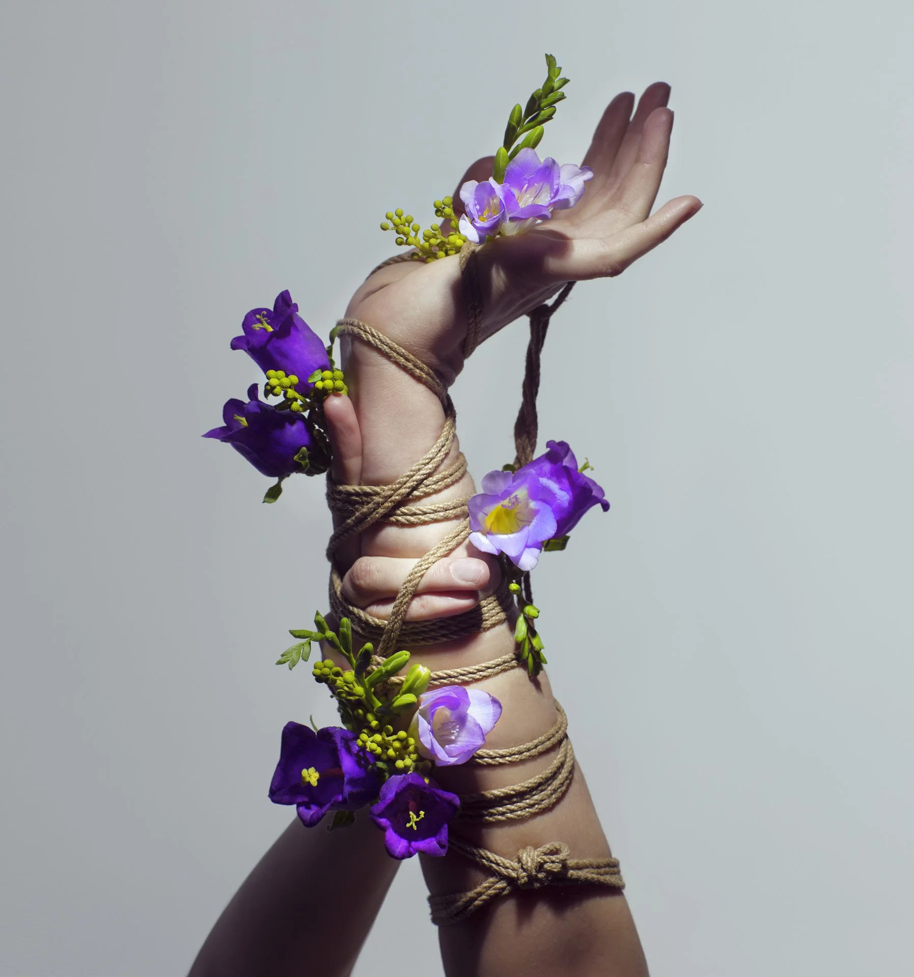 A hand with fingers outstretched is wrapped with beige ropes and decorated with purple and yellow flowers and green buds, against a plain light gray background.