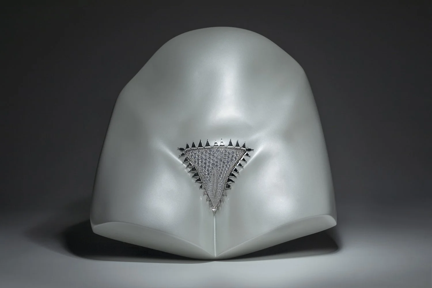 A white, abstract sculpture with a diamond-studded triangular emblem in the center, set against a dark background.