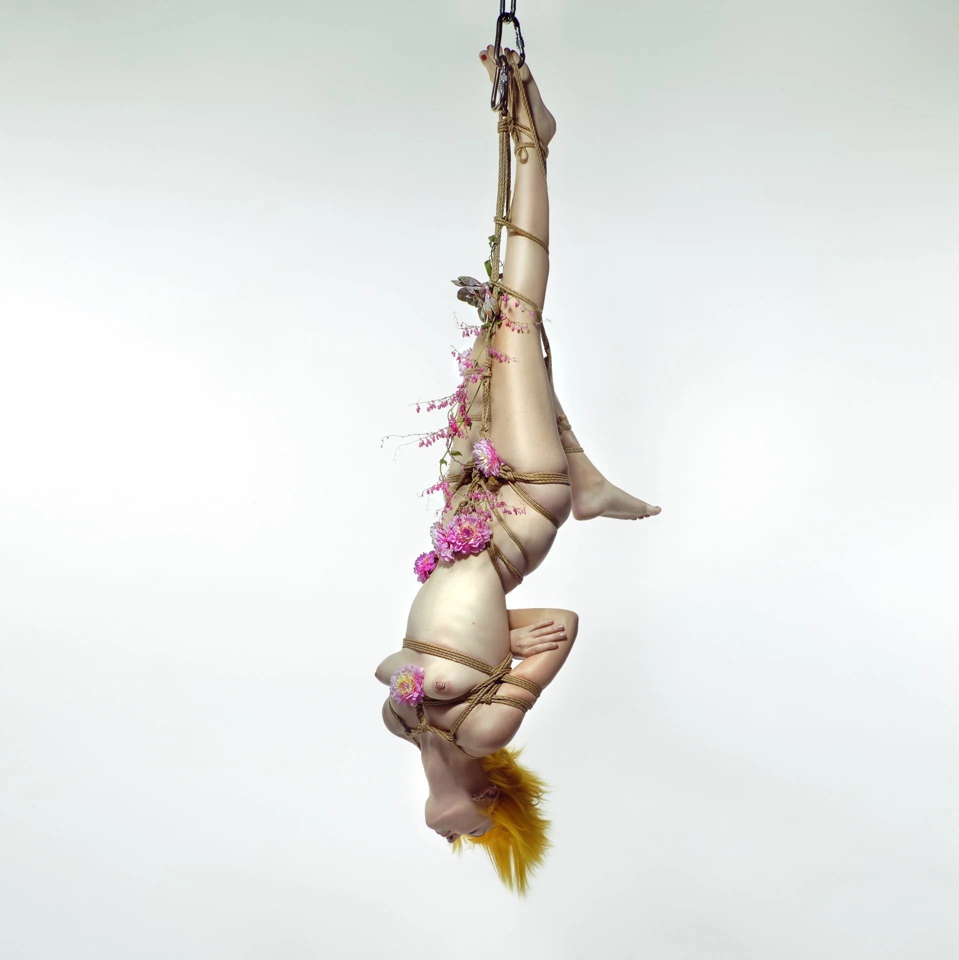 A person with orange hair is hanging upside down from a rope, adorned with pink flowers and green foliage, against a plain white background.