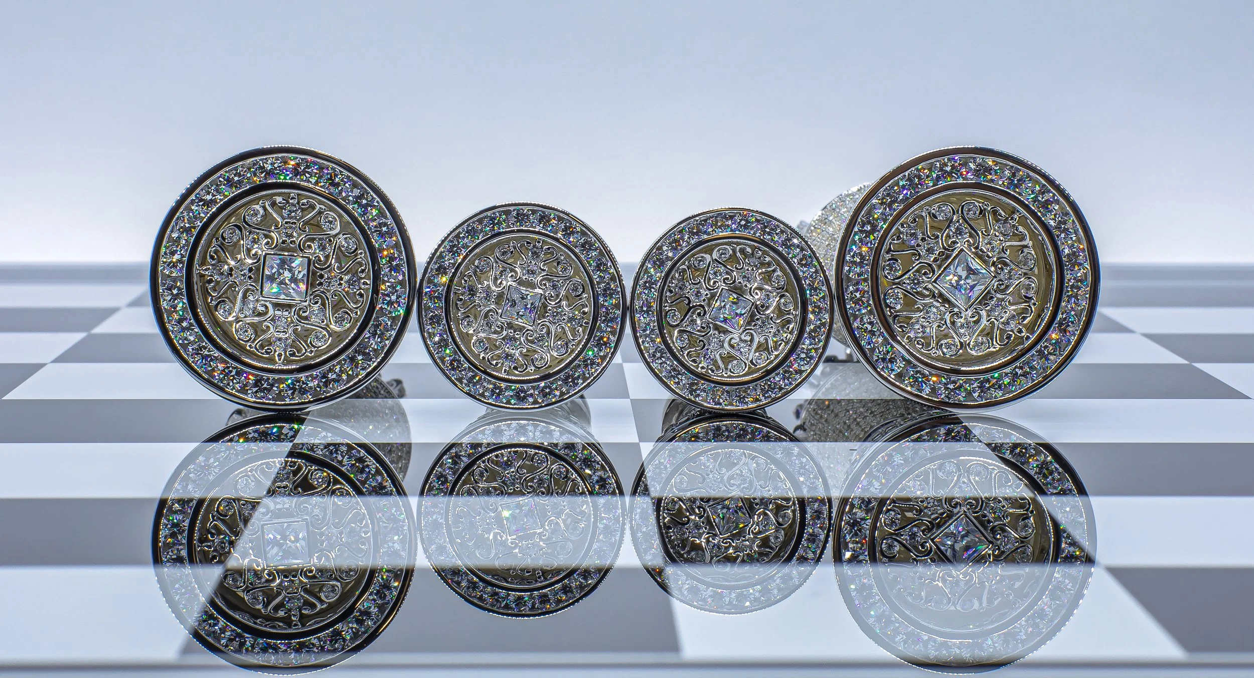Four decorative circular objects with intricate designs and embedded sparkling stones, placed on a black and white checkered surface with their reflections visible below.