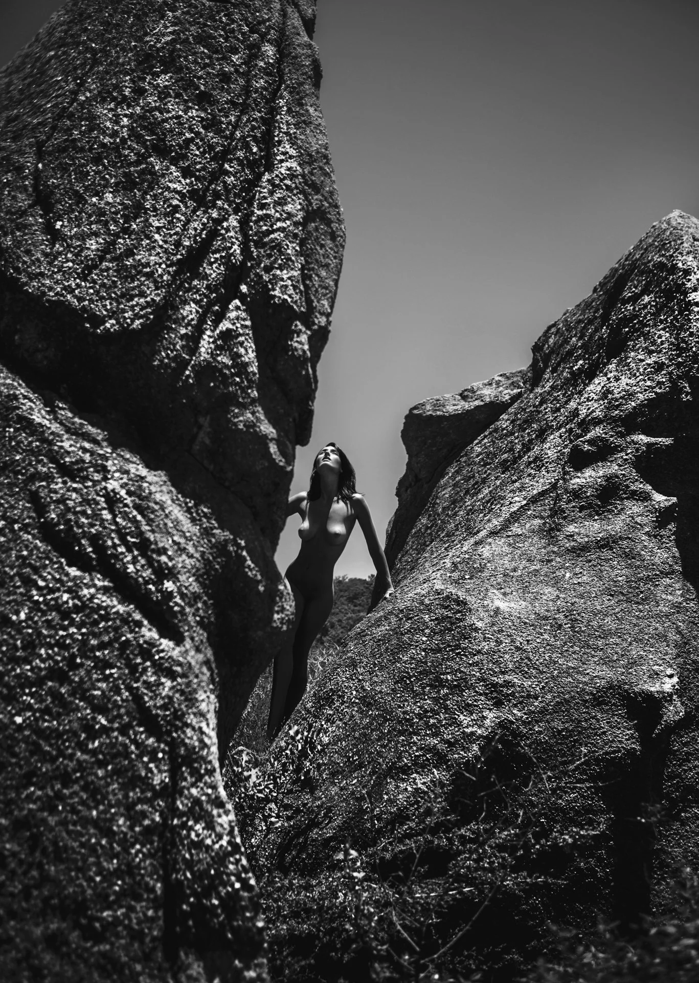 A woman standing nude between large, textured rocks outdoors, looking up towards the sky.