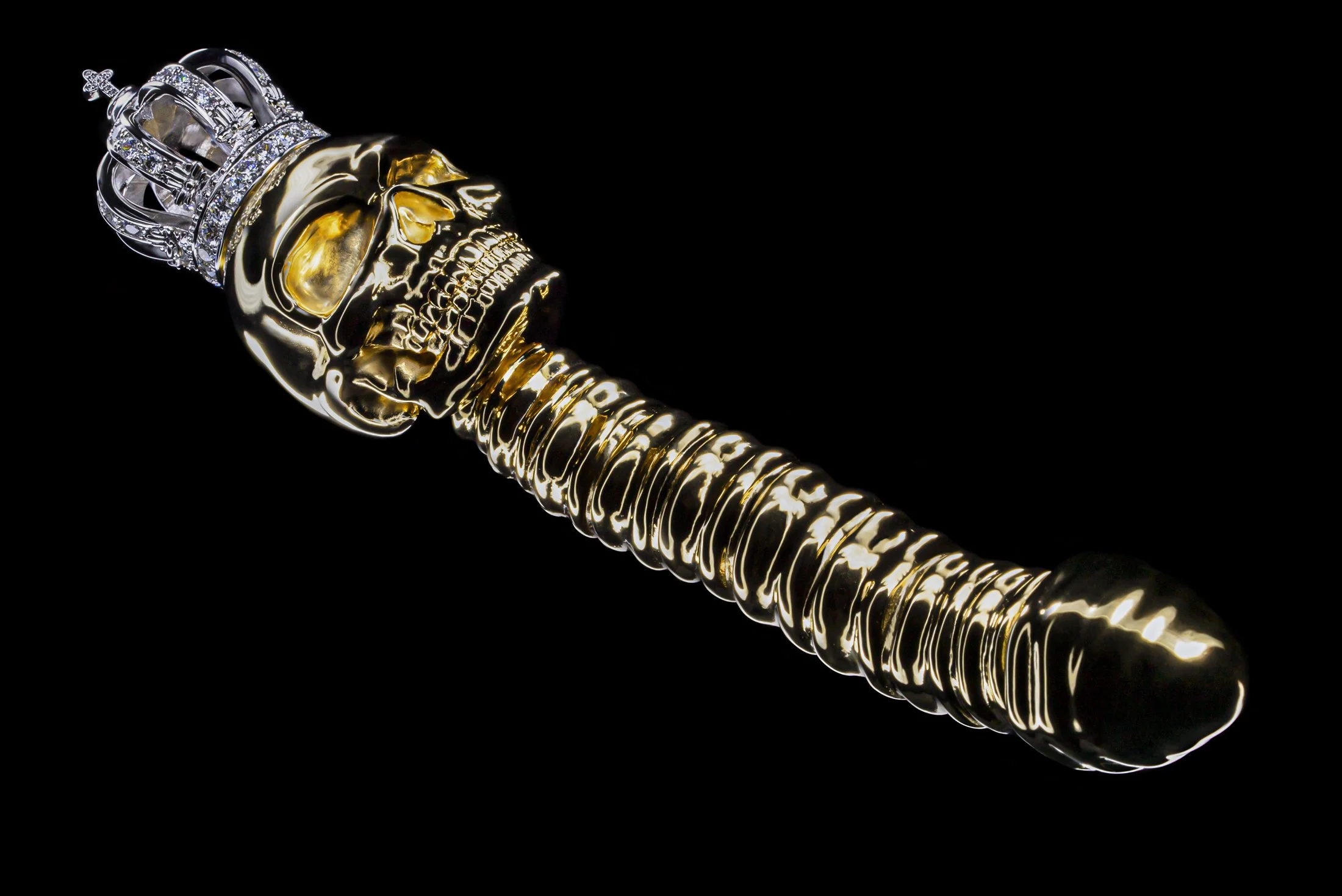 A decorative object resembling a metallic skull with gold and silver accents, topped with a silver crown adorned with diamonds, set against a black background.