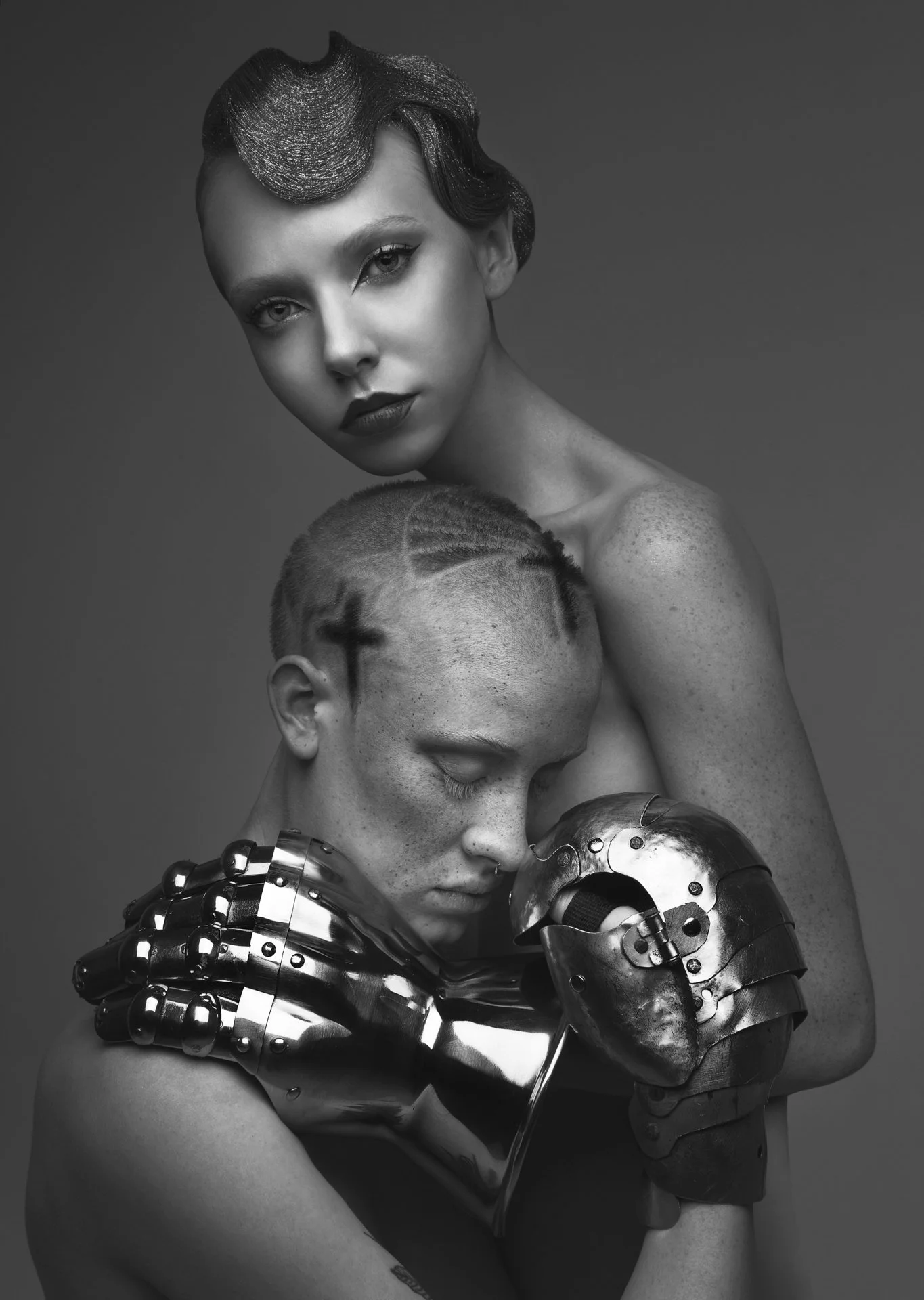 Black and white photo of a woman with vintage hairstyle and makeup, holding a man with shaved head and freckled skin, wearing a metallic robotic glove and arm, both with serious expressions.