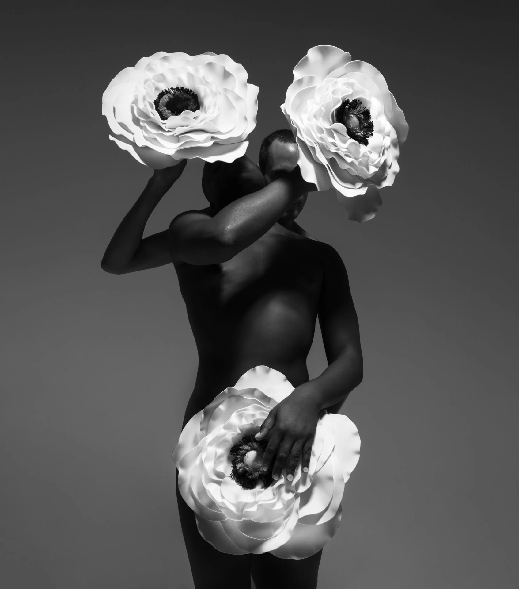 A black-and-white photo of a nude person holding large artificial white flowers over their head and covering their private areas. The person is turne in a profile view against a plain background.