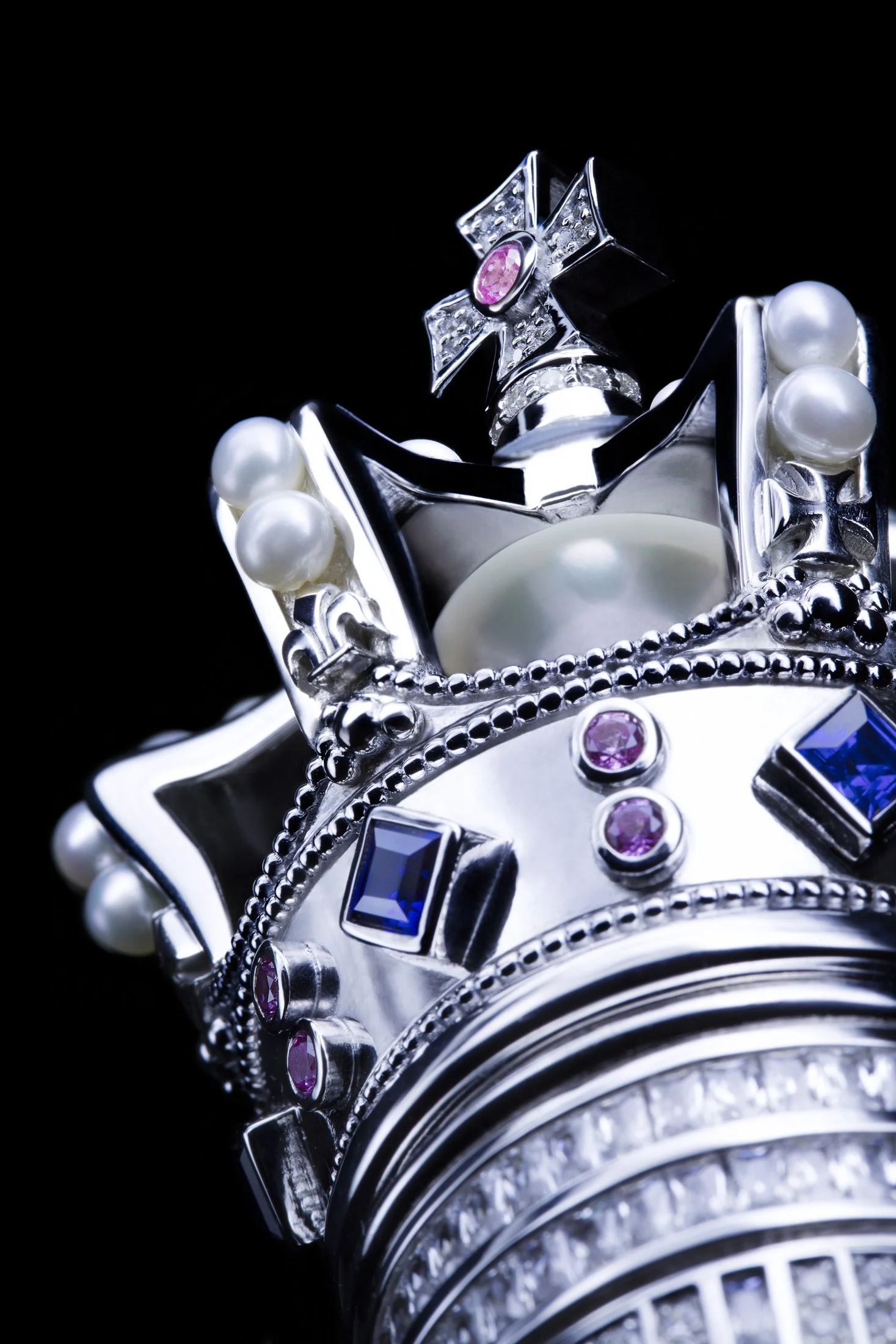 Close-up of ornate silver jewelry with pearls, sapphires, pink gemstones, and a cross on a black background.