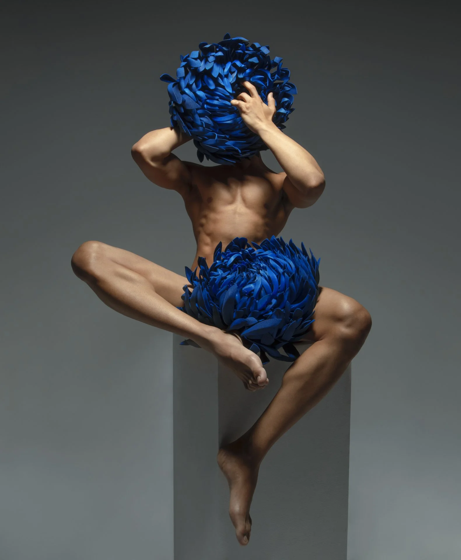 A muscular person sitting cross-legged on a gray pedestal, holding large blue flower-like structures over their head and around their waist in a contemporary art pose.