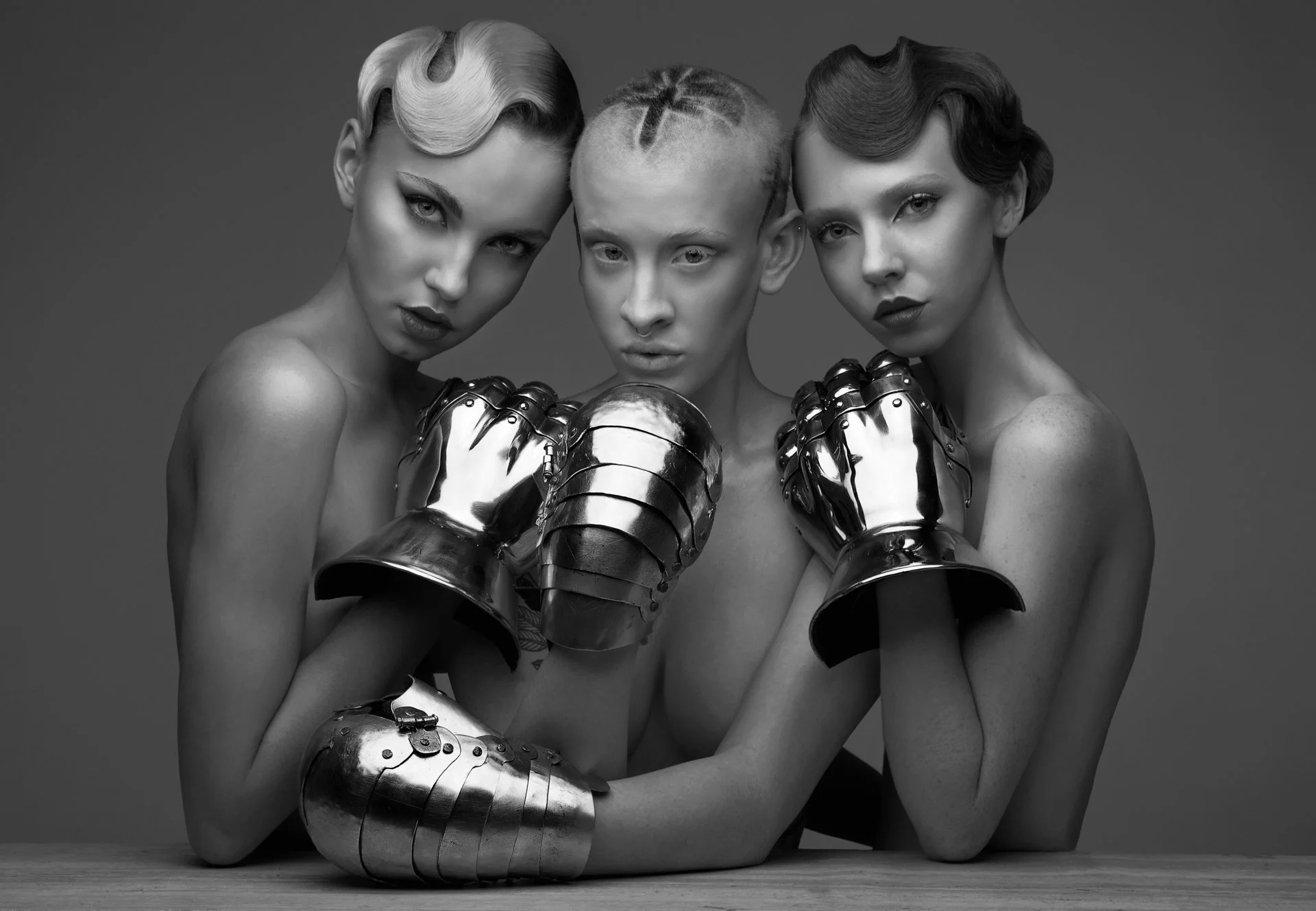 Black and white photo of three topless women with short, styled hair, wearing metallic arm and hand armor, looking at the camera with intense expressions.