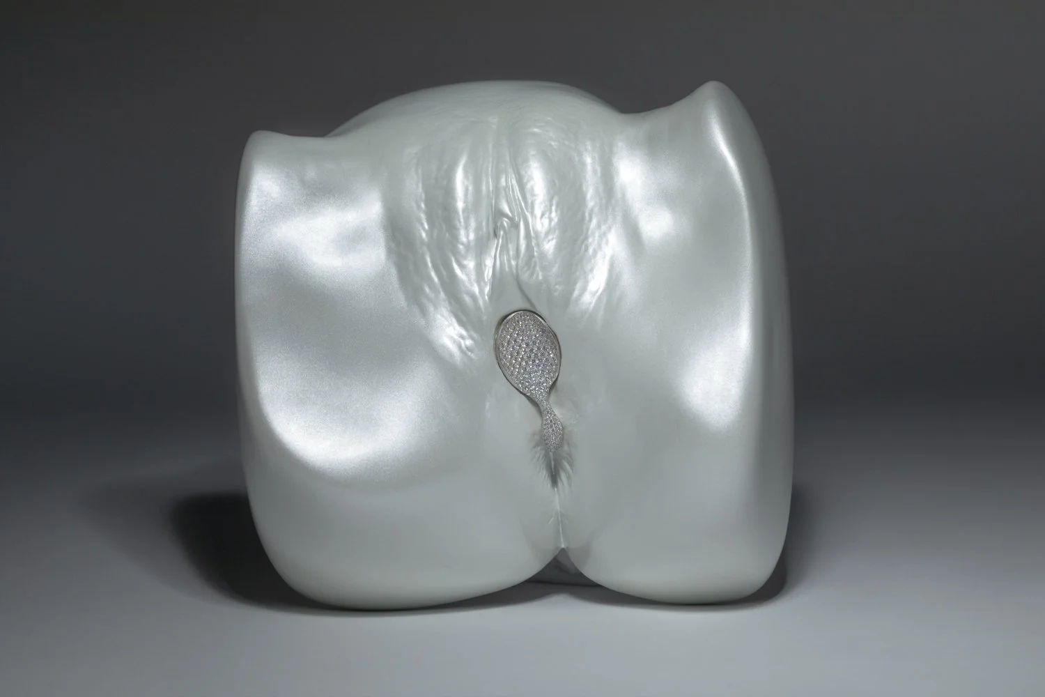 A silver, glossy sculpture resembling a human buttocks with a small, textured, drip-shaped object in the center.
