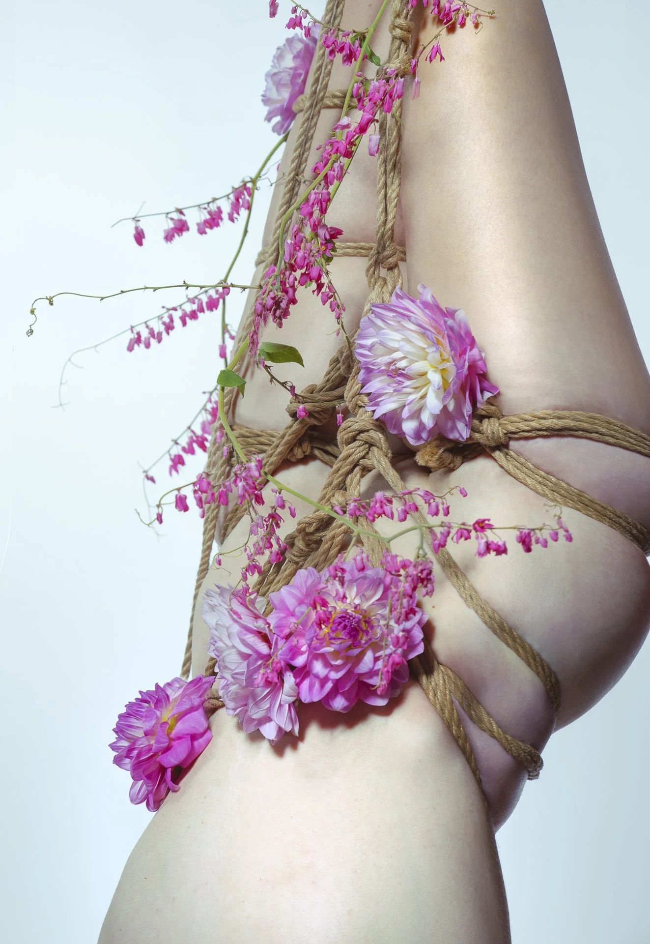 Close-up of a nude person's body wrapped with brown rope, adorned with pink and white flowers.